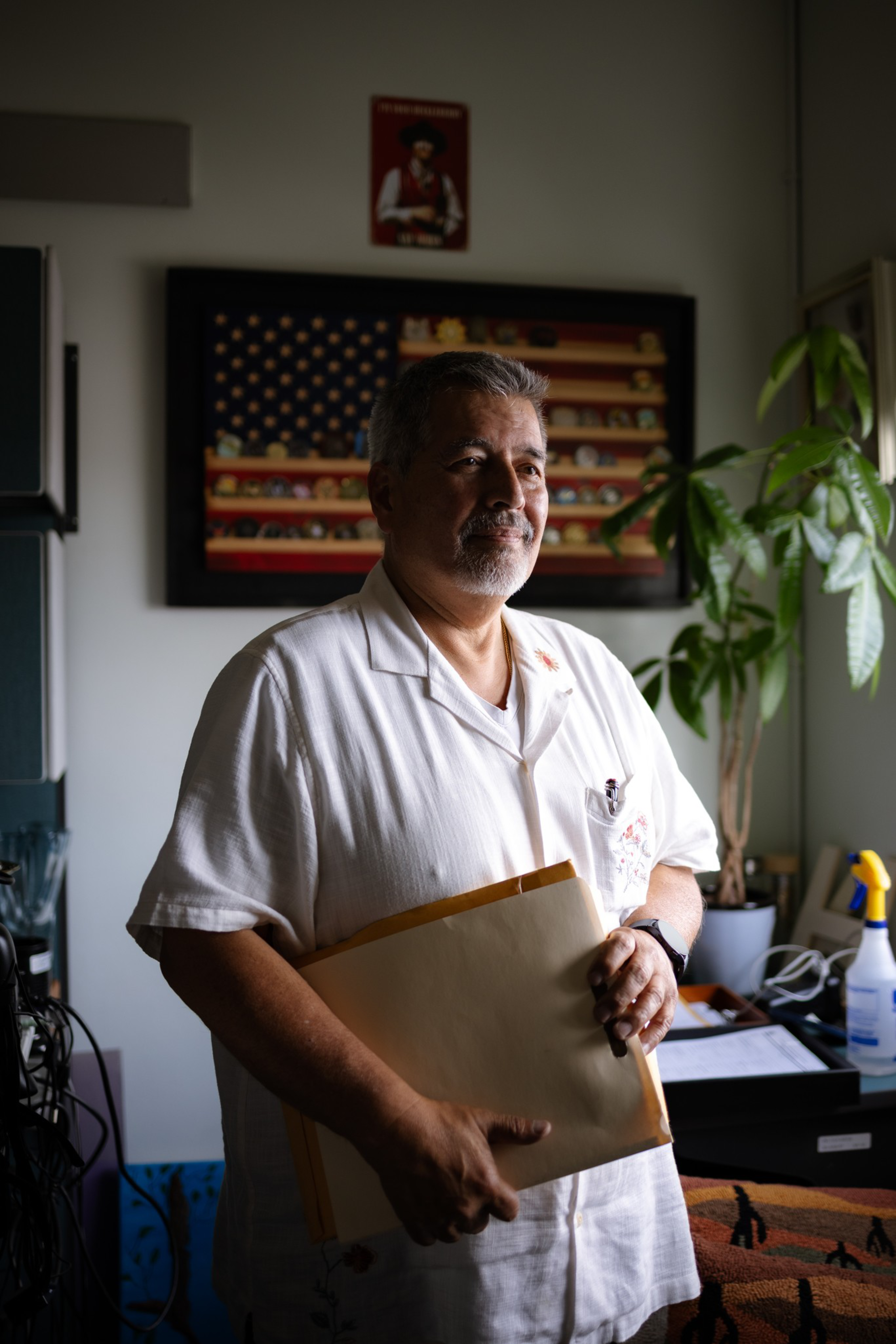 A man in a white shirt holds a large envelope, standing in a room with a framed American flag and a tall plant in the background.
