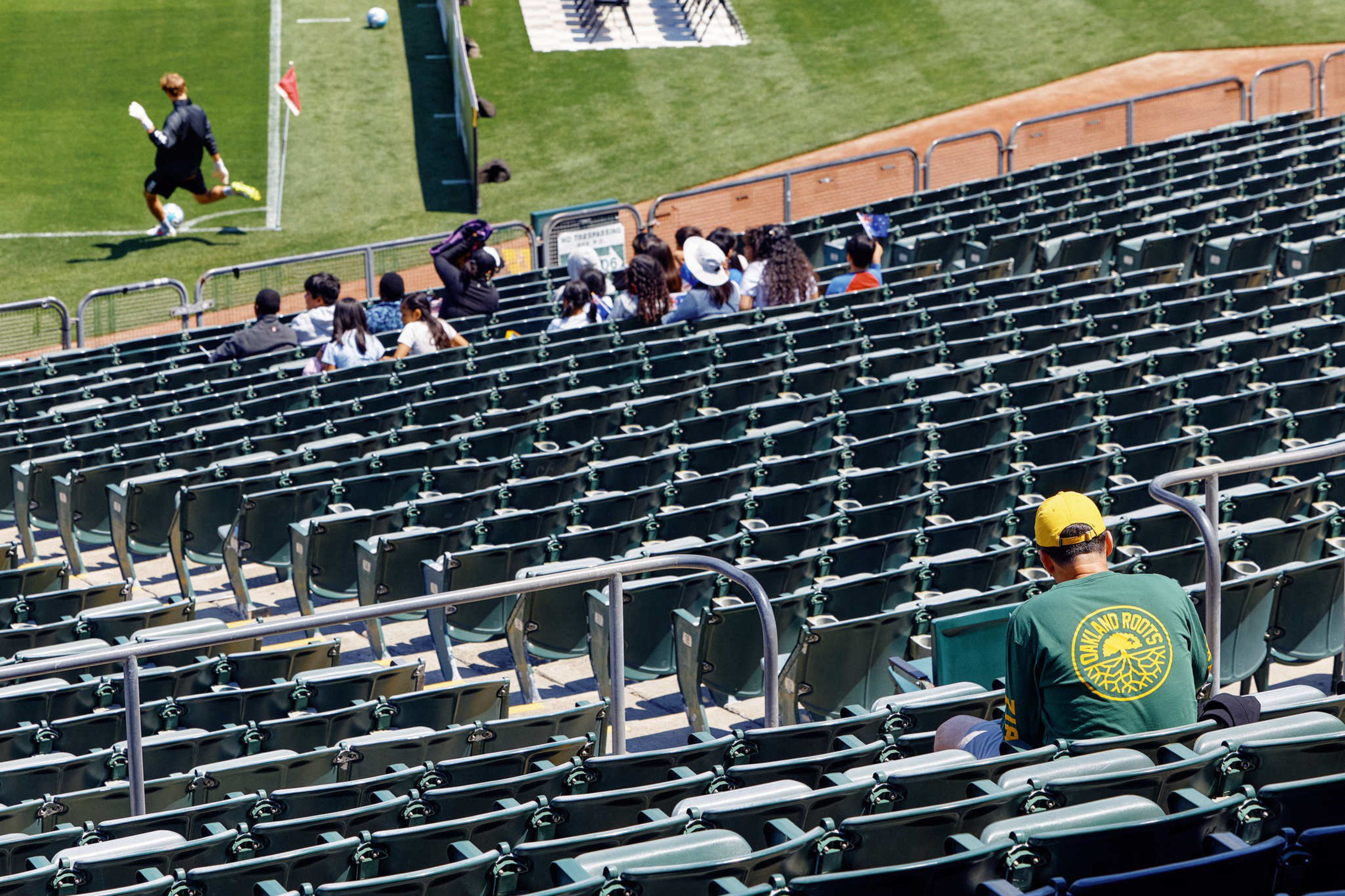 A nearly empty stadium with green seats shows a lone person in an Oakland Roots hoodie sitting while a group watches a soccer player near the goal.