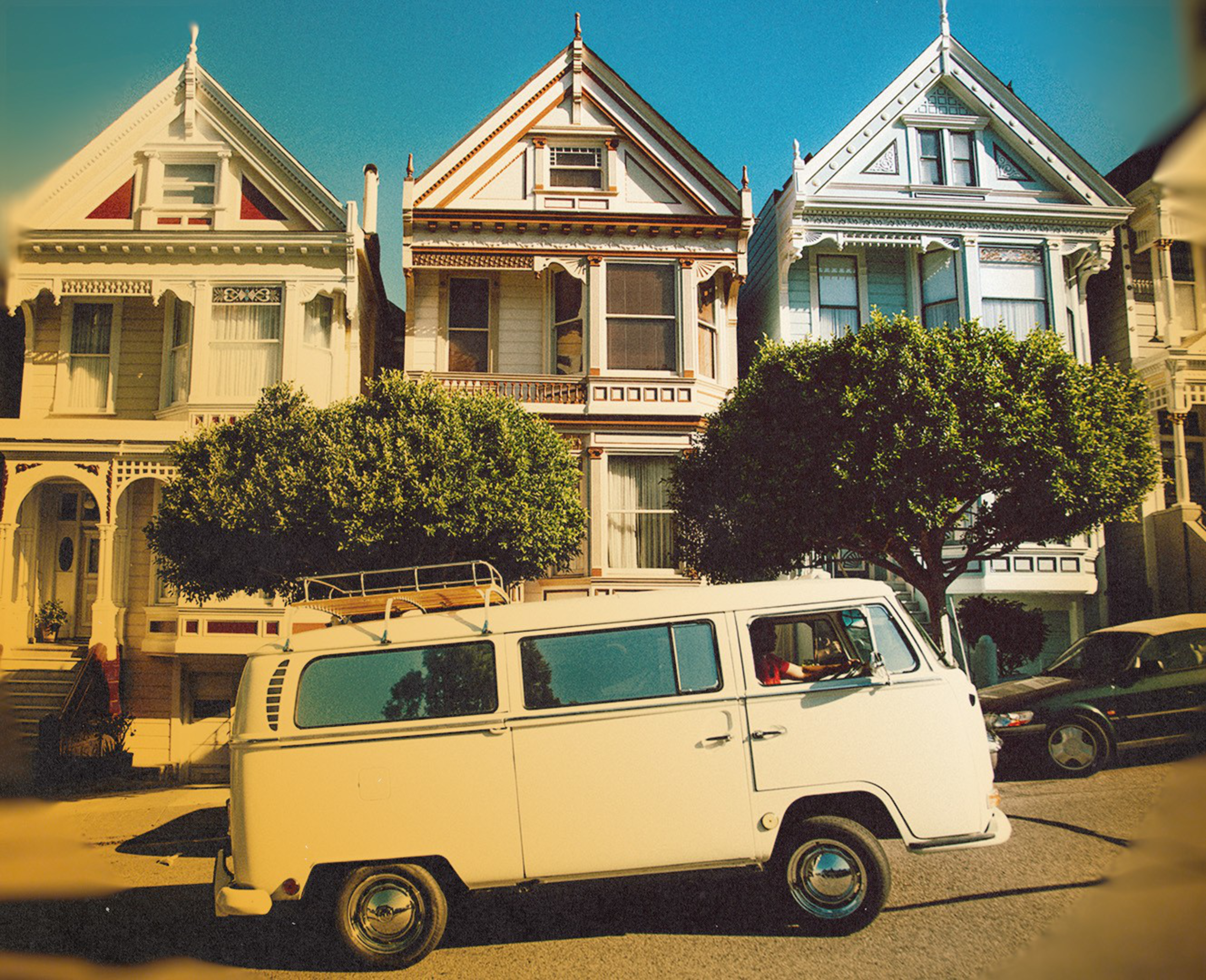 A vintage white van is parked in front of three colorful Victorian-style houses with detailed trim and two leafy trees on the sunny street.