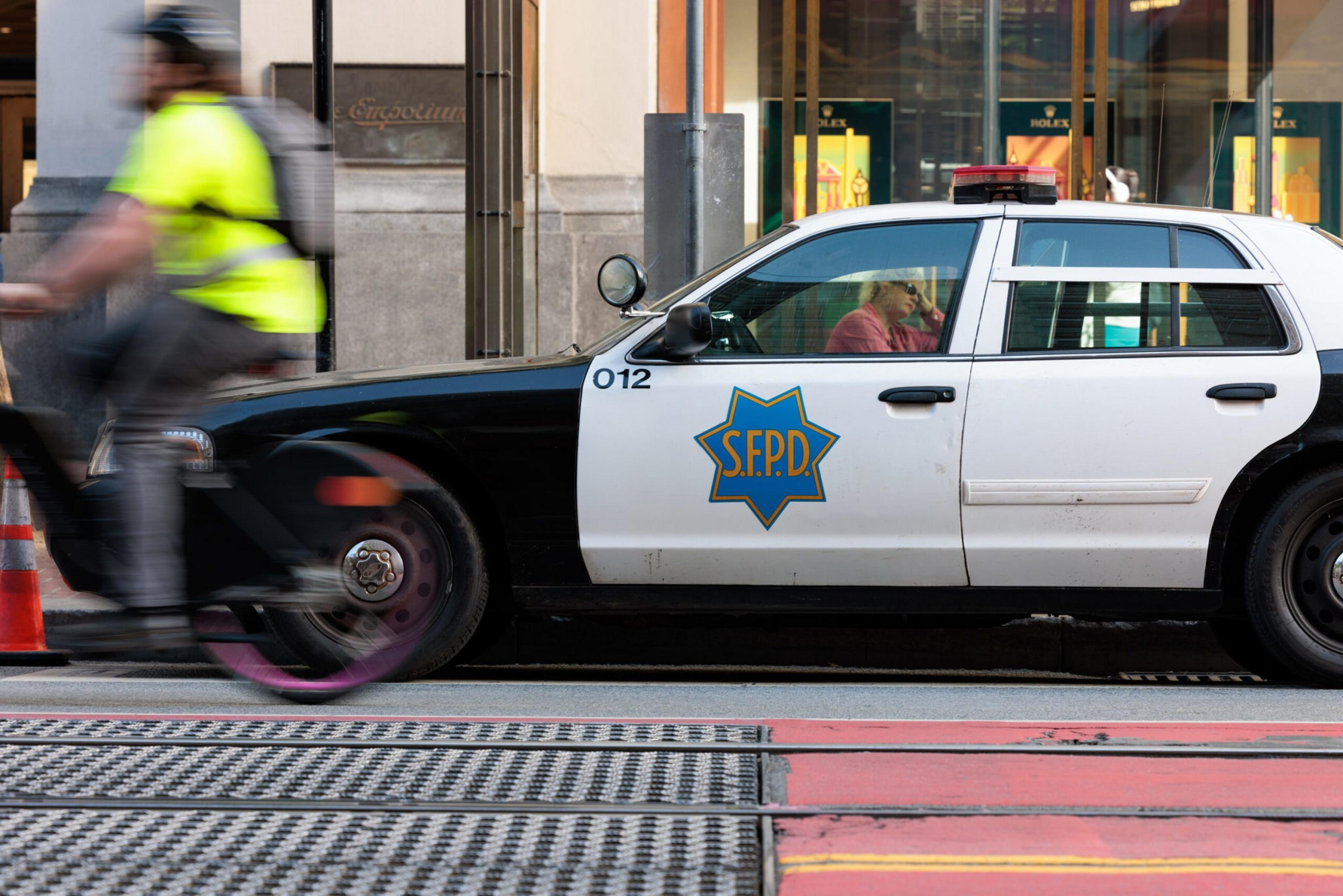A police car marked 'SFPD' is parked as a blurred cyclist in a neon vest rides by.