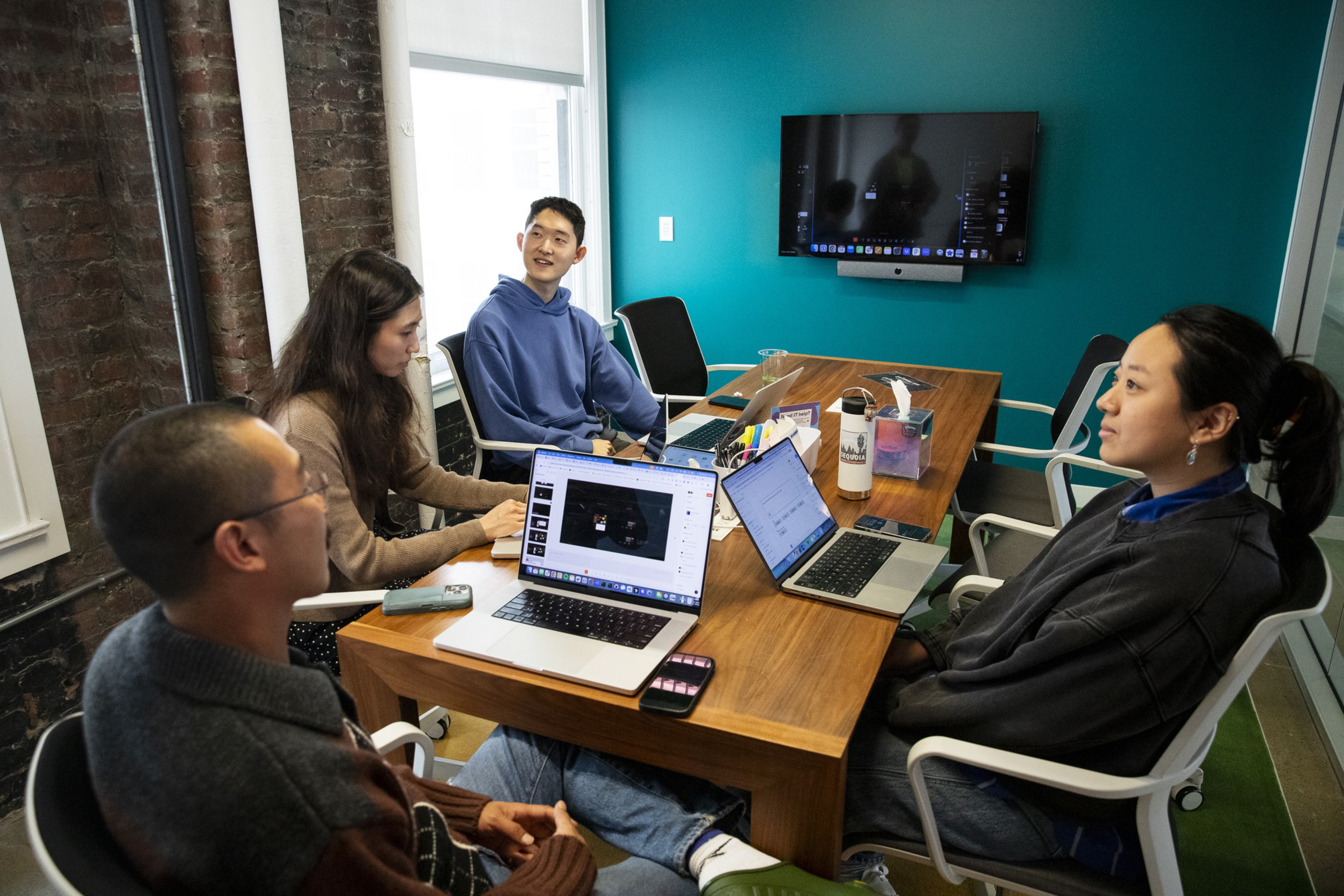 Four people sit around a wooden table in a meeting room, each with a laptop, engaged in discussion, with a screen mounted on a teal wall behind them.