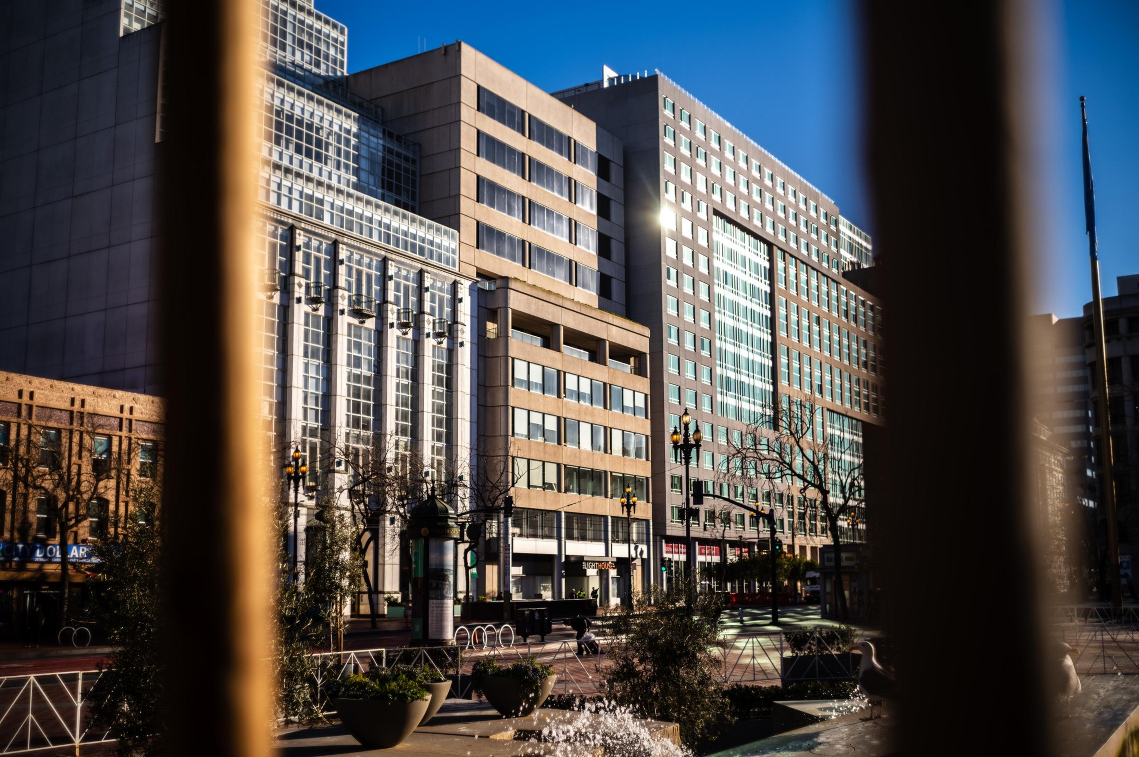 Urban scene with modern buildings, clear blue sky, street lamps, and a few trees, viewed through a blurred foreground.