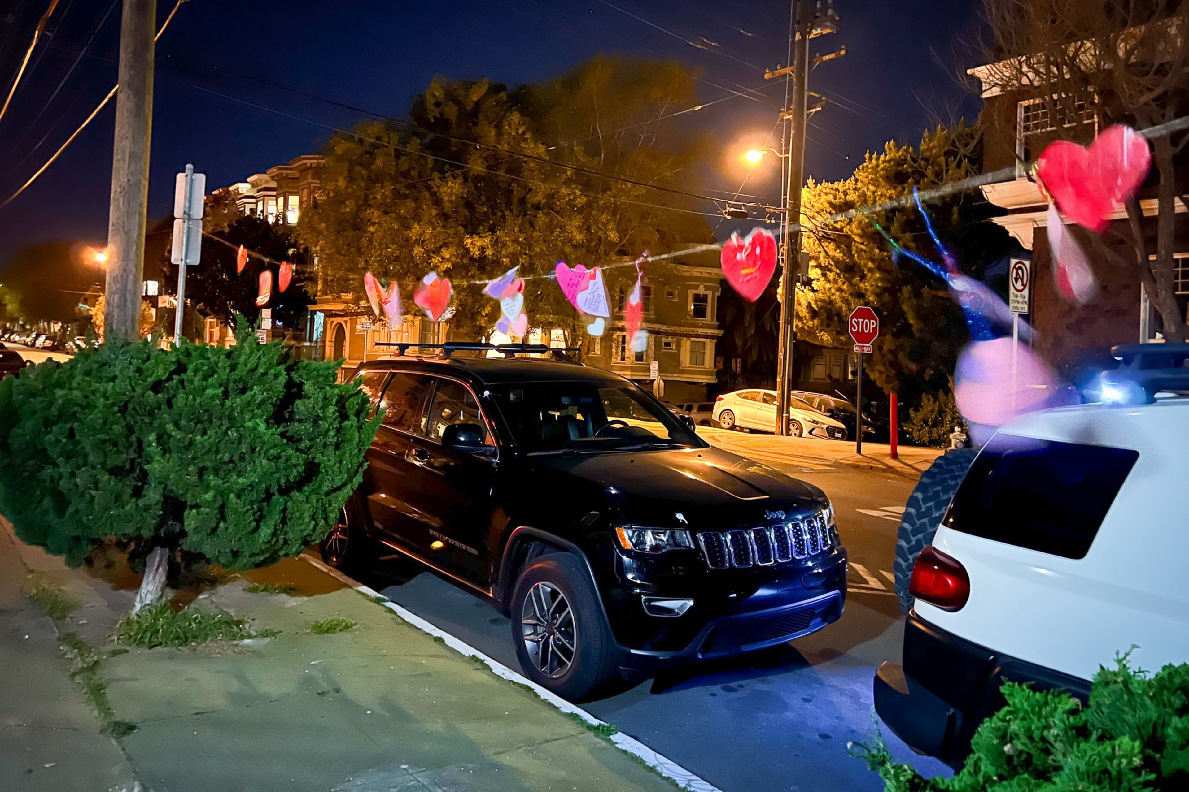 A nighttime street scene features a black SUV and a white car parked along the curb with red heart-shaped decorations strung between them.