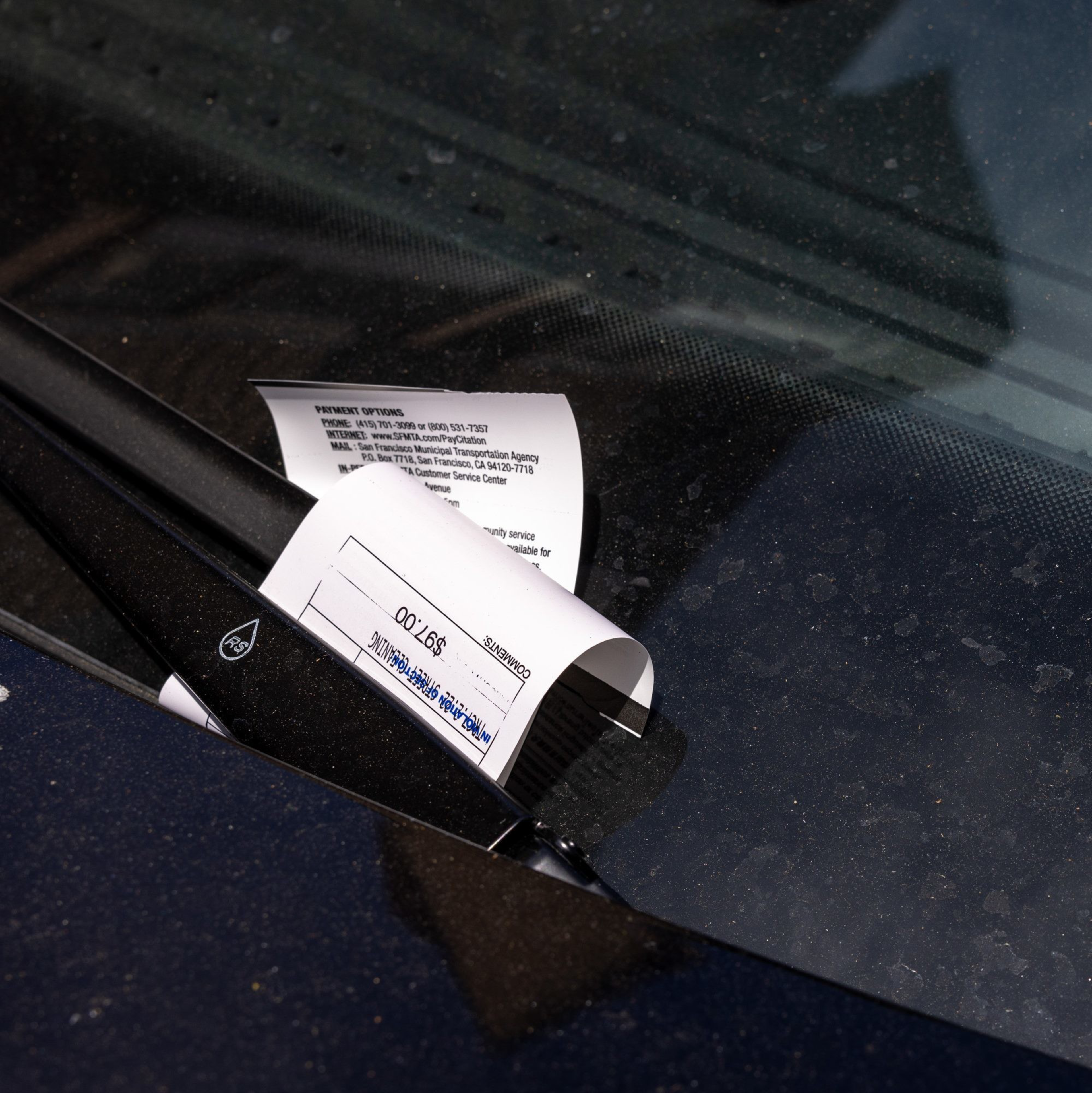 A parking ticket is placed under the wiper of a dark car, visible on a dusty windshield.