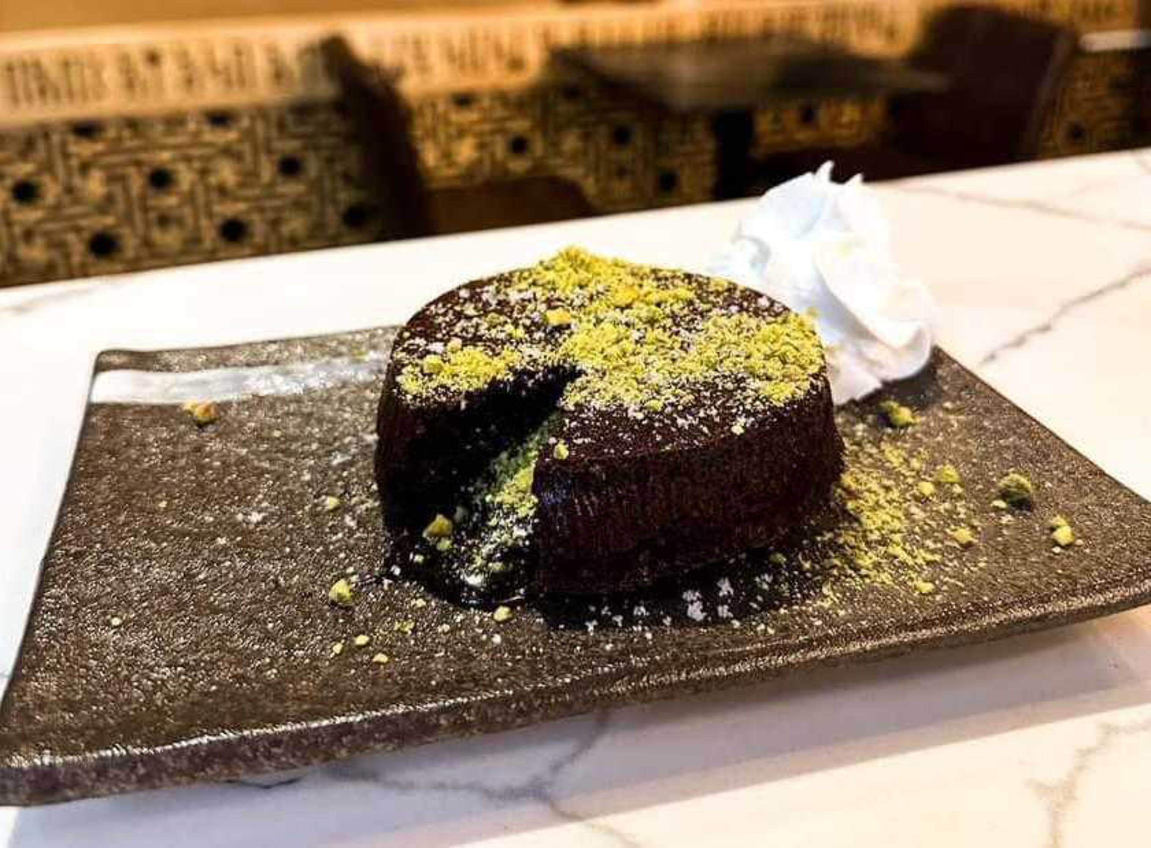 A chocolate lava cake with a gooey center, topped with crushed pistachios and powdered sugar, served with a dollop of whipped cream on a black rectangular plate.