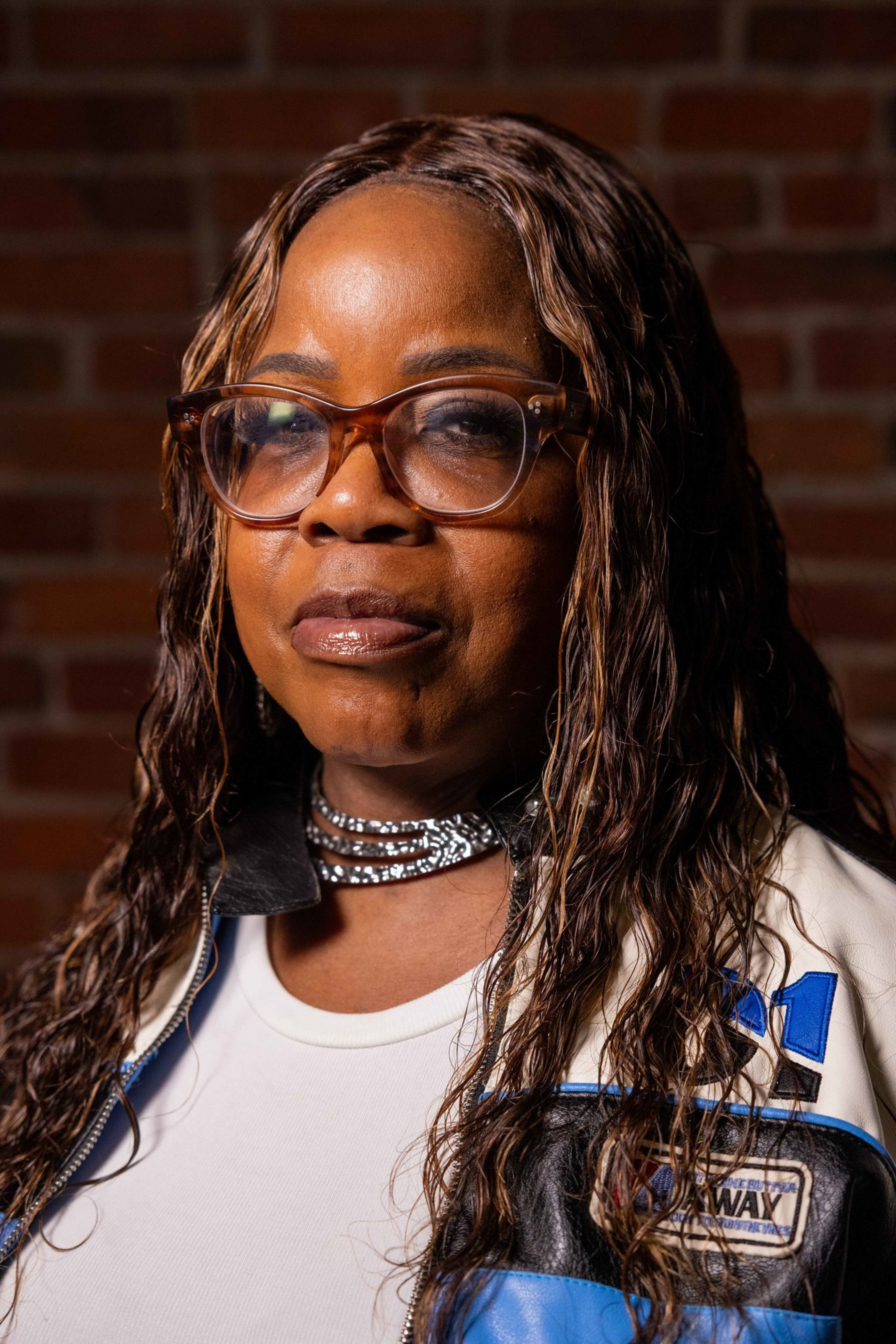A woman with long, wavy hair wears large glasses, a white shirt, a blue and black jacket, and a shiny choker necklace against a brick wall background.