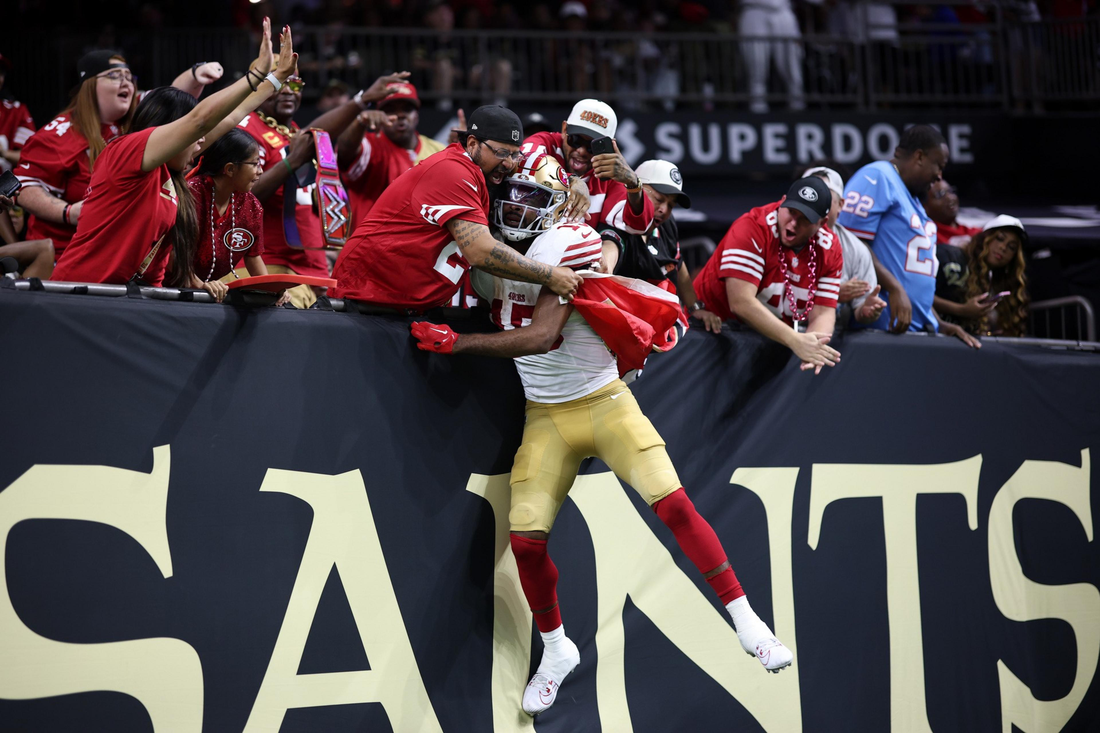 A football player in a white and gold uniform jumps to embrace fans wearing red jerseys over a barrier with “SAINTS” written on it.