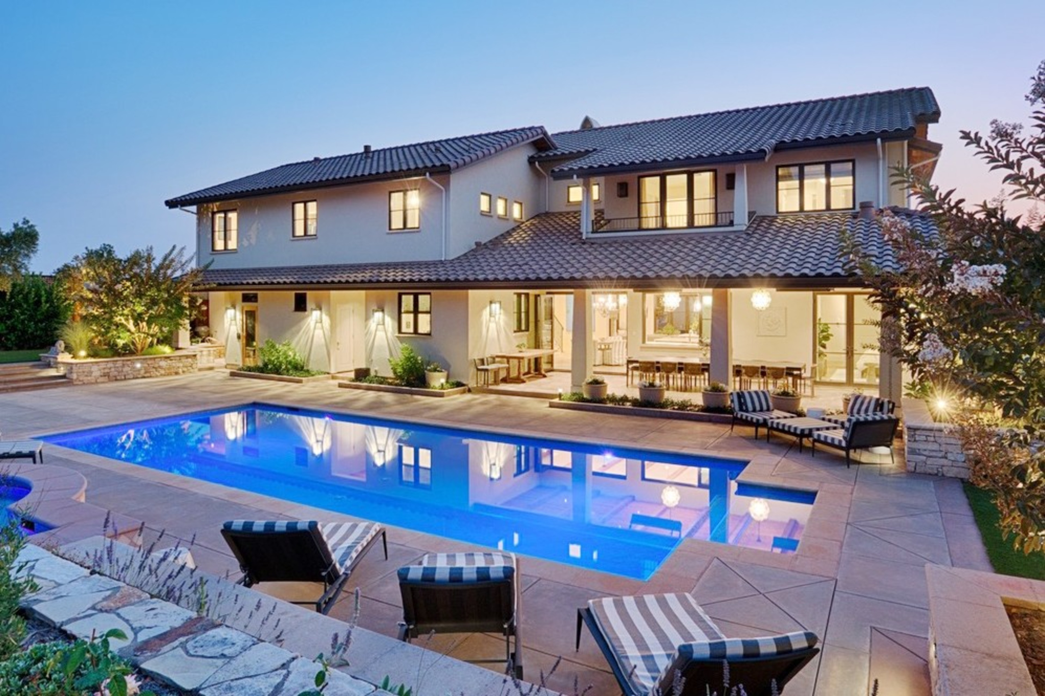 A luxurious two-story house with a tiled roof features a spacious backyard with a clear blue swimming pool, patio furniture, and well-maintained landscaping at dusk.