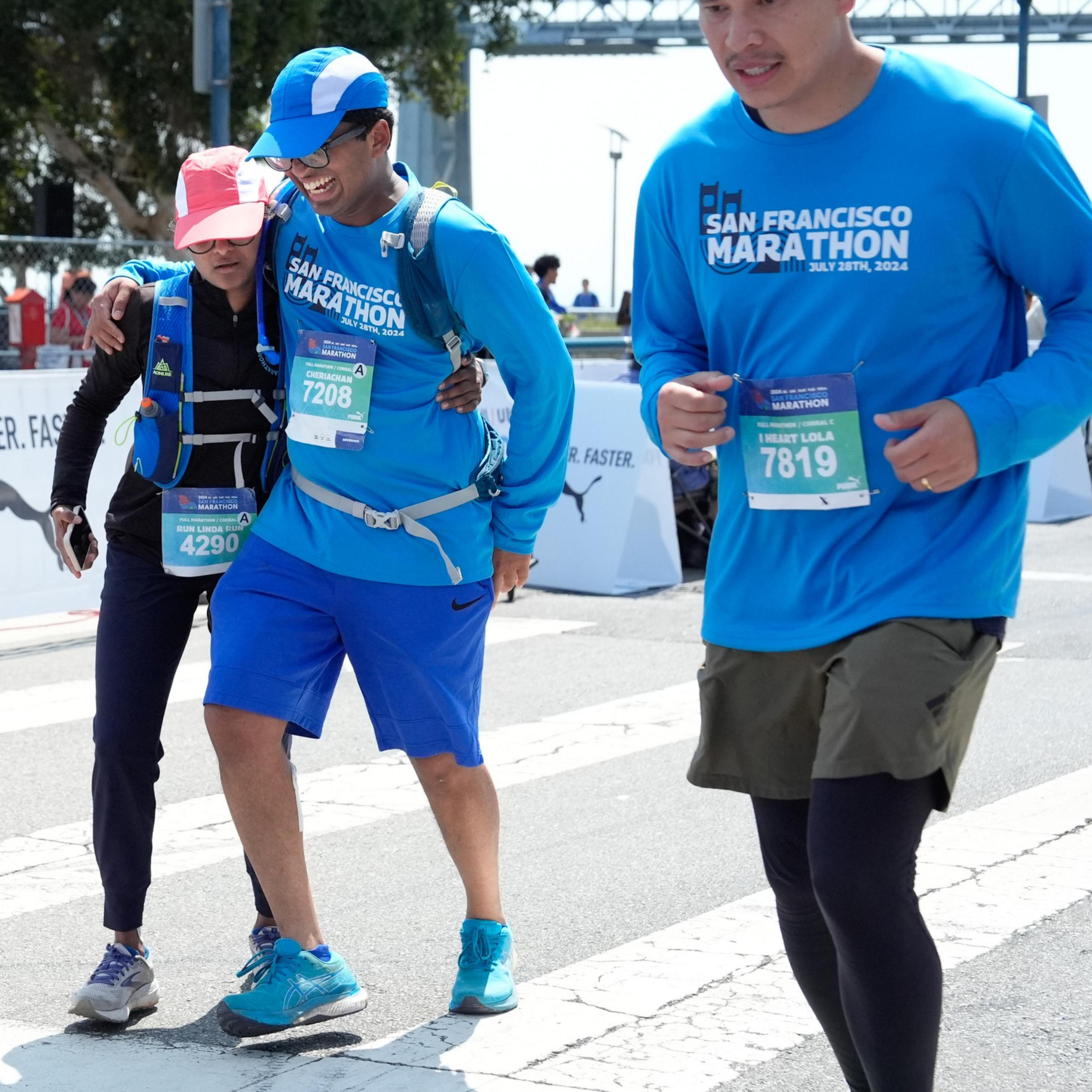 Three runners wearing blue “San Francisco Marathon” shirts cross the finish line together, two of them assisting each other while smiling.