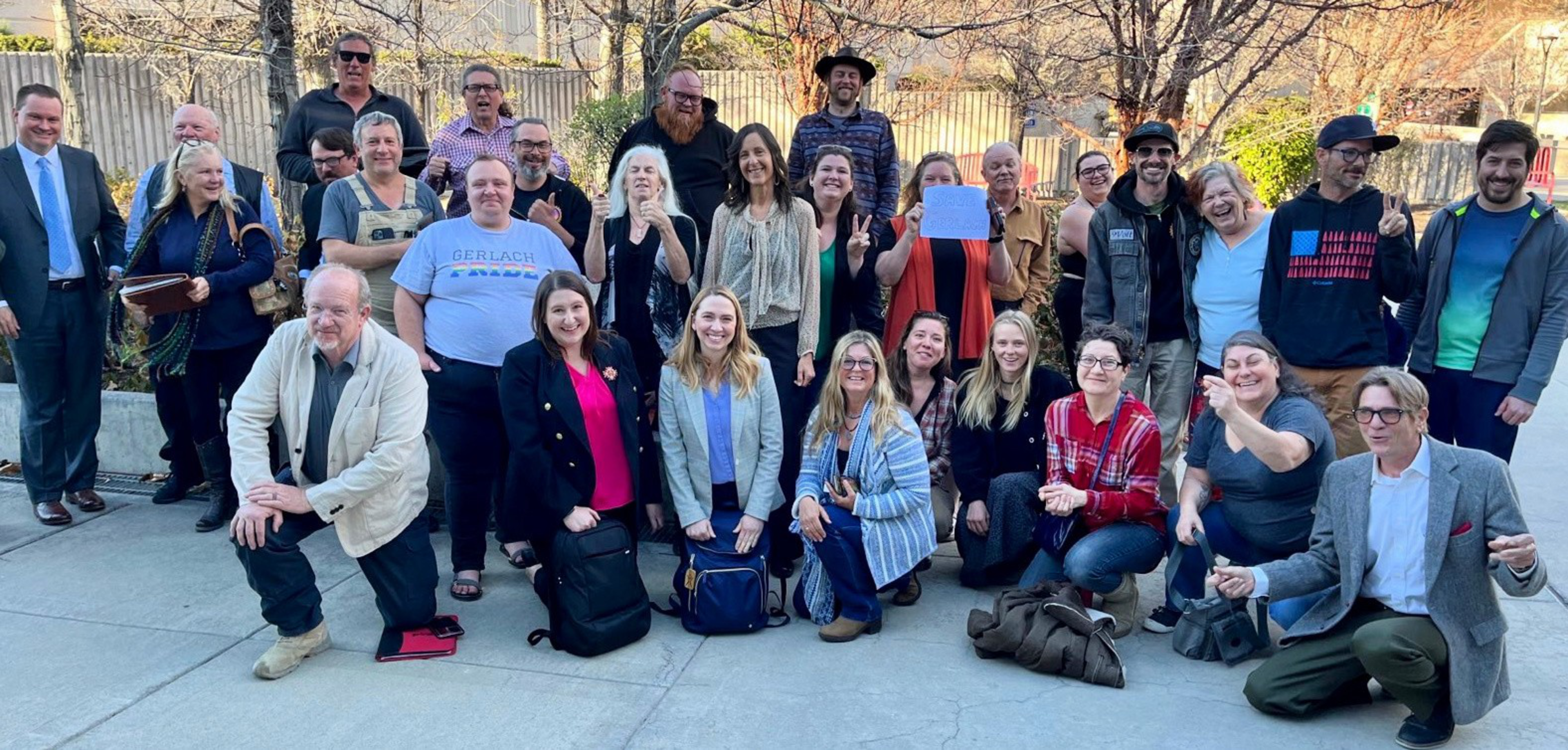 A diverse group of men and women, some standing and some kneeling, pose outdoors on concrete with trees and a fence behind them.