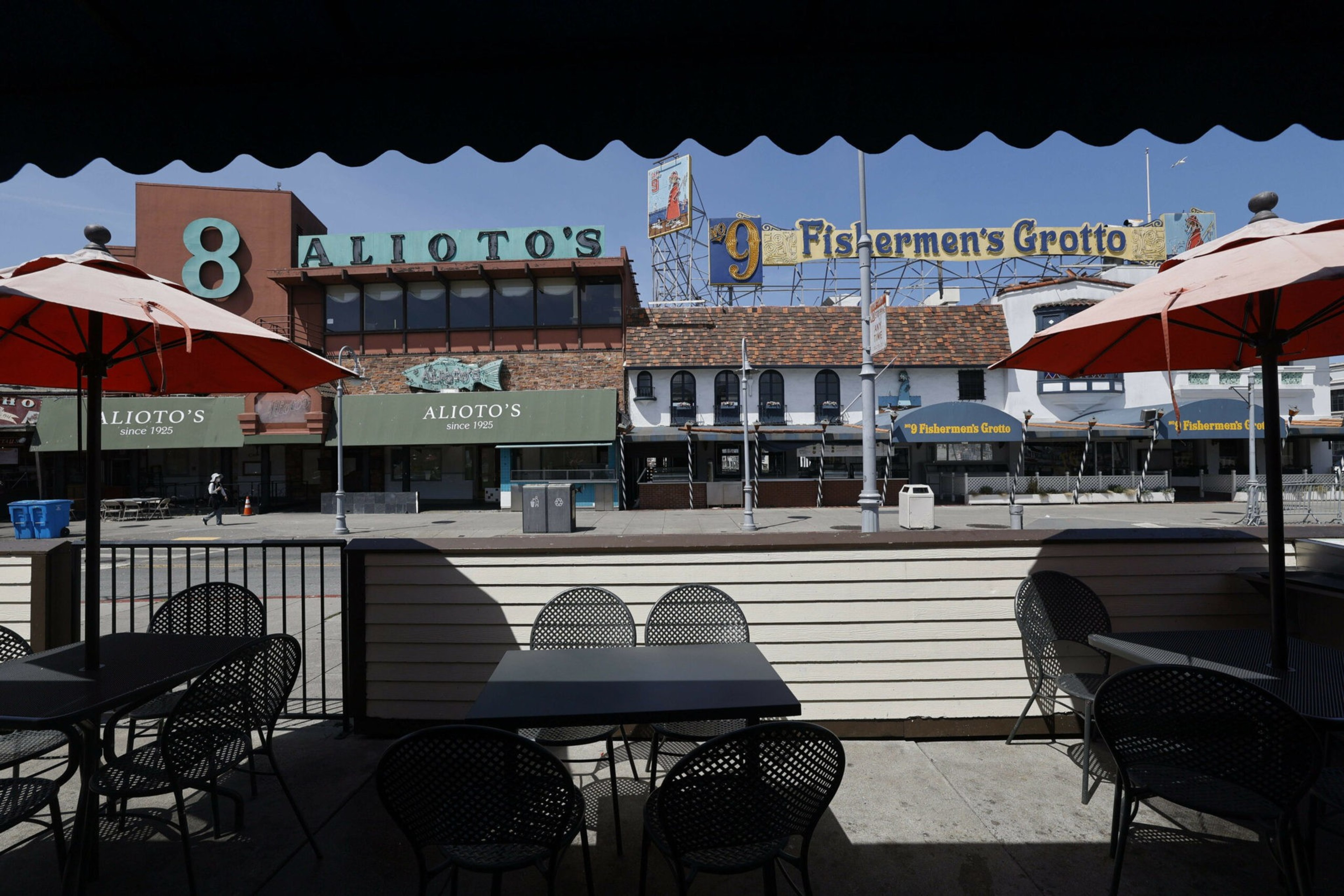 Outdoor seating with red umbrellas faces across the street to the buildings of Alioto's and Fishermen's Grotto restaurants under a clear blue sky.