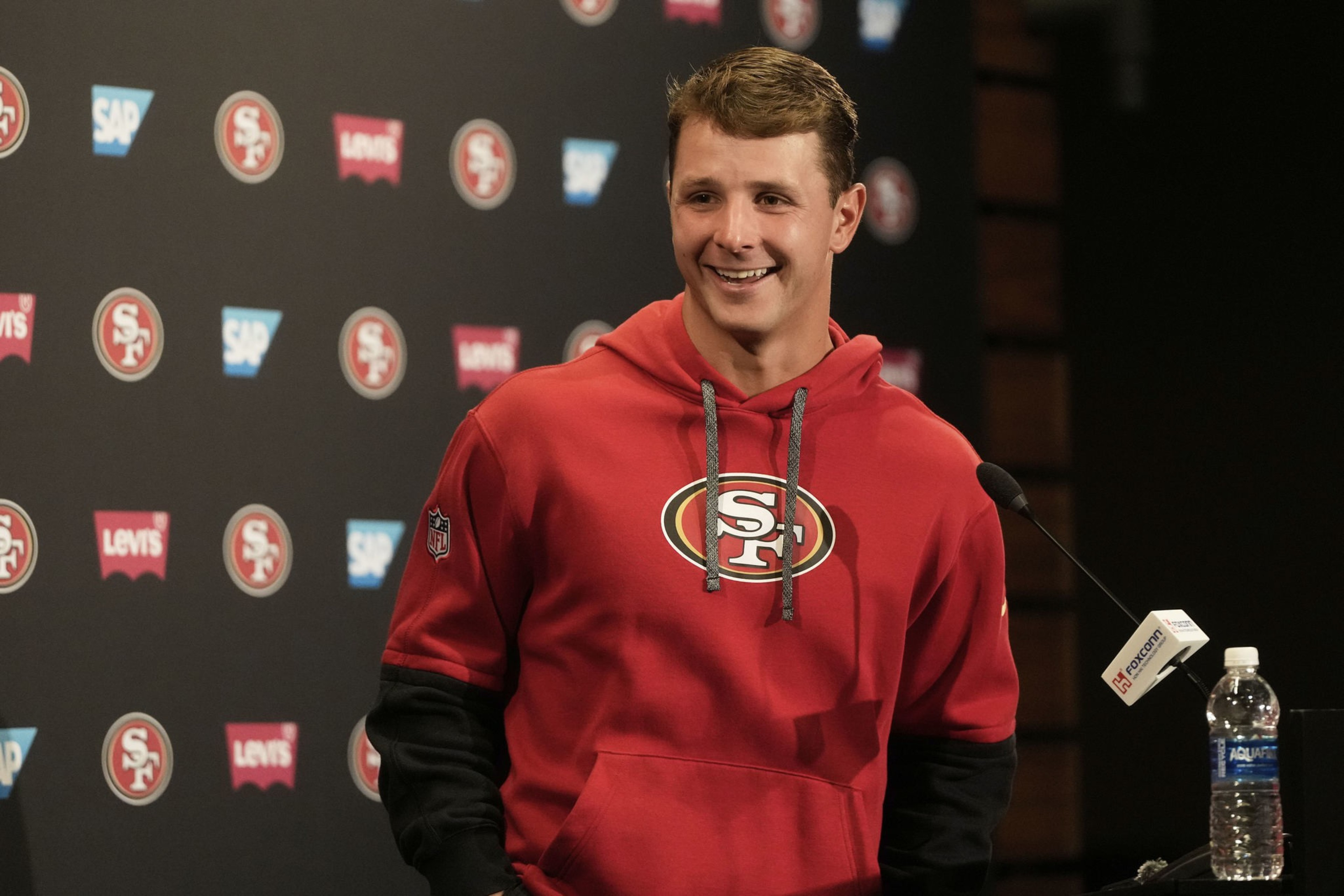 A smiling man in a red 49ers hoodie stands at a podium with microphones, in front of a branded backdrop featuring SAP and Levi's logos.