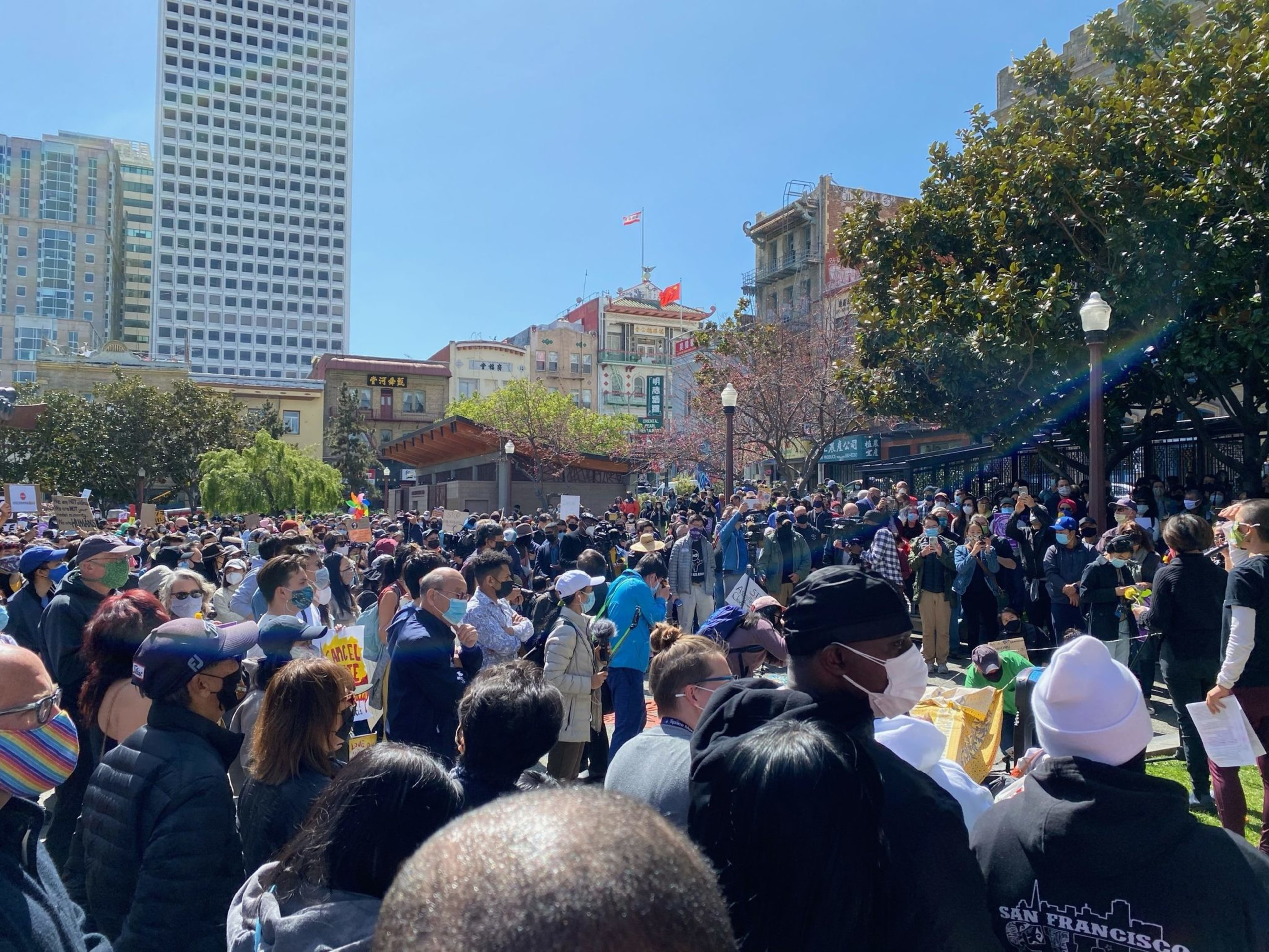 A large crowd of people wearing masks gathers outdoors in a city park, surrounded by trees and tall buildings under a clear sky.