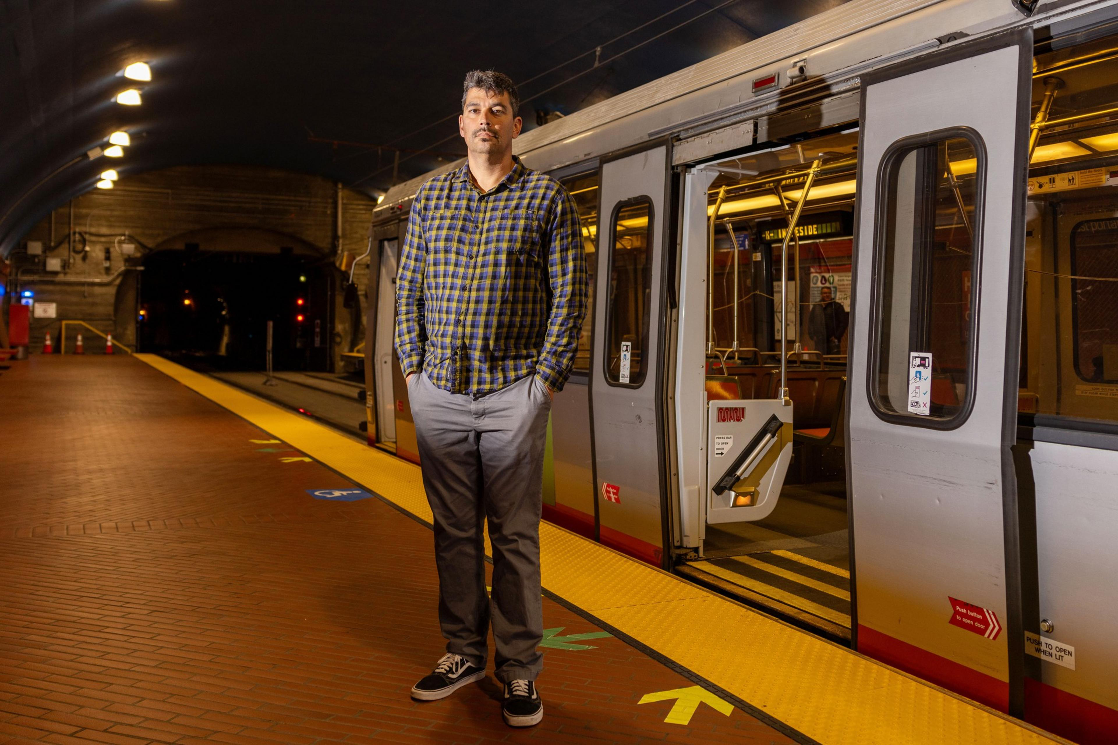A man in a plaid shirt and gray pants stands on a well-lit subway platform next to an open subway car door. The platform is empty and clean with yellow safety guides.