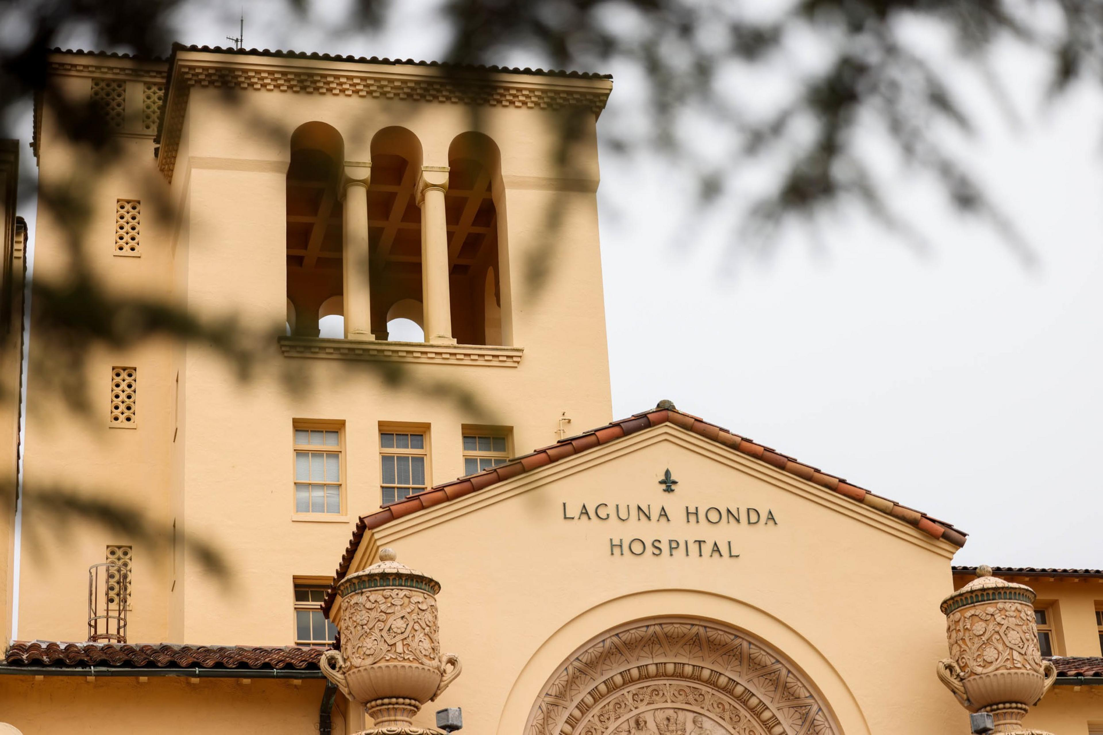 A beige building with Spanish-style architecture houses Laguna Honda Hospital, featuring terracotta roof tiles and ornate columns.