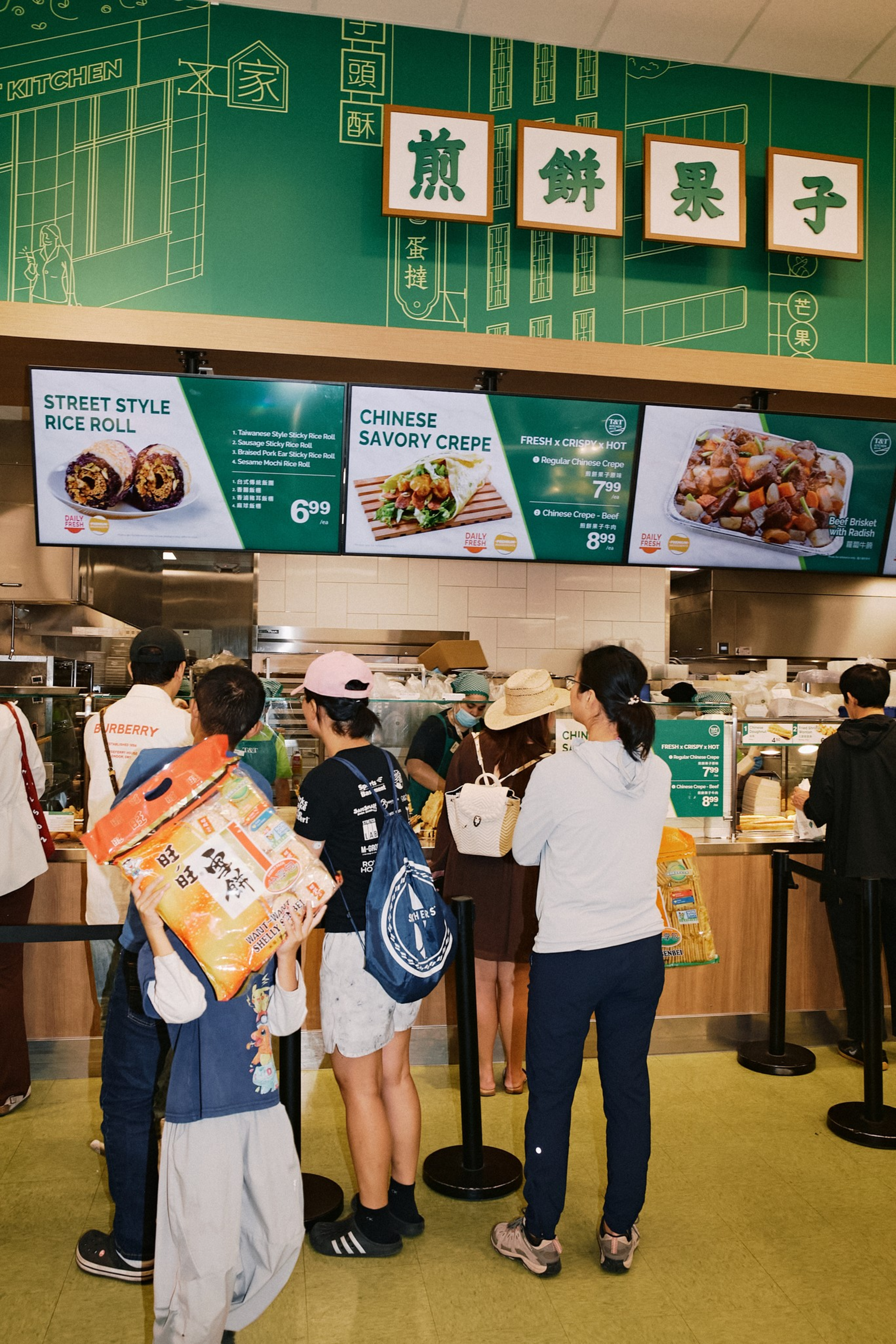 Customers stand in line ordering food at a Chinese kitchen counter, with menu screens displaying rice rolls, savory crepes, and brisket dishes.