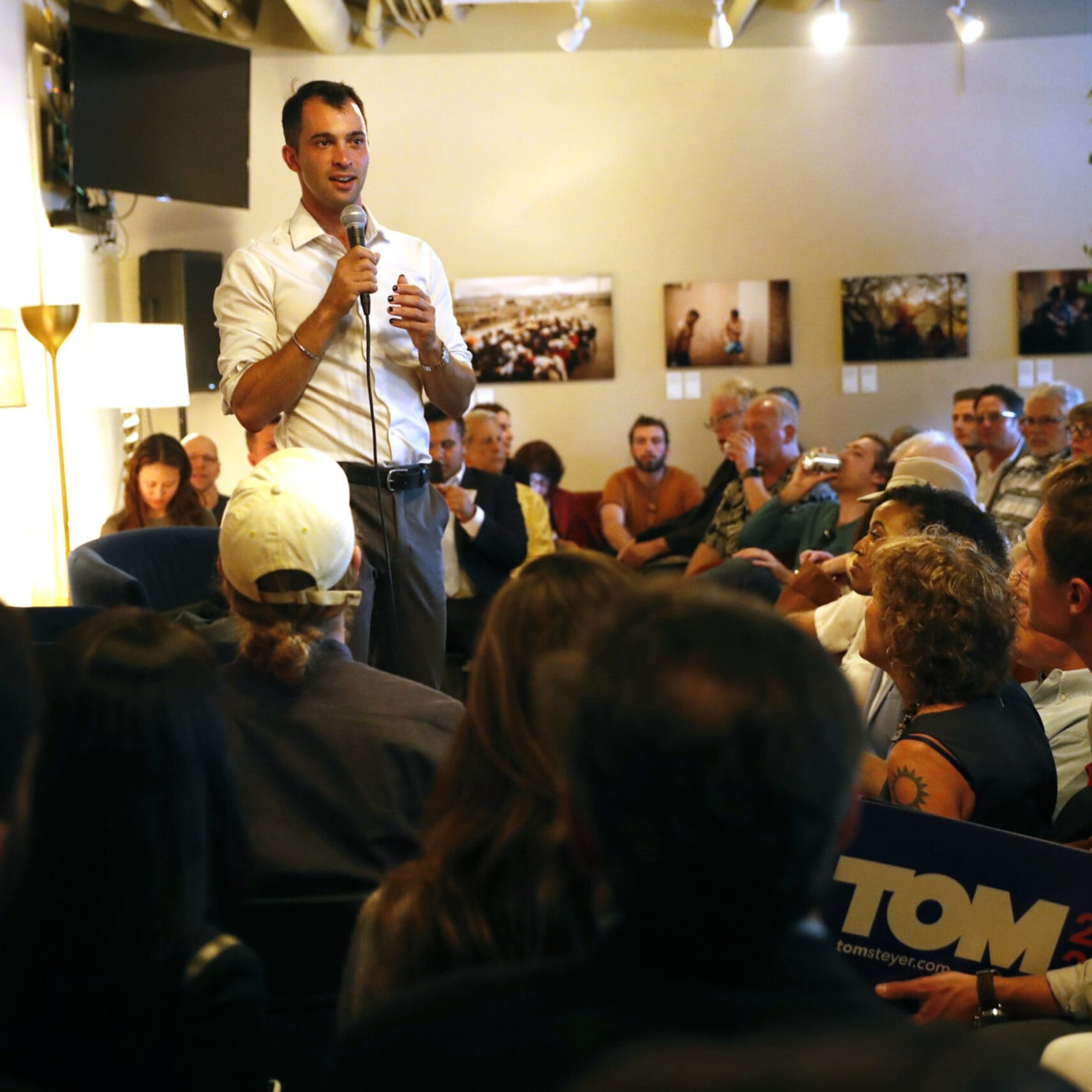 A man in a white shirt speaks with a microphone to a seated, attentive audience in a warmly lit room decorated with photos and plants.