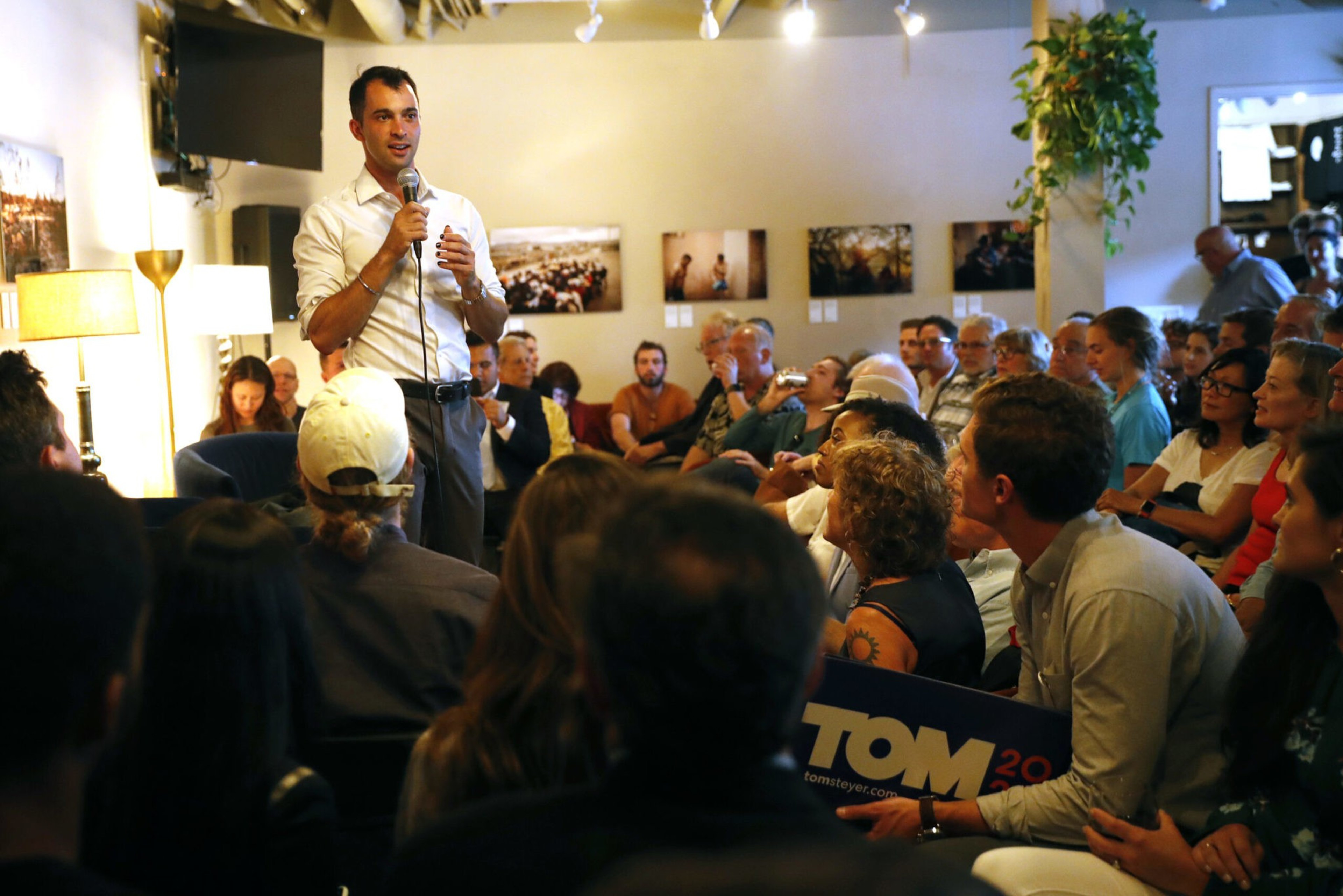 A man in a white shirt speaks with a microphone to a seated, attentive audience in a warmly lit room decorated with photos and plants.