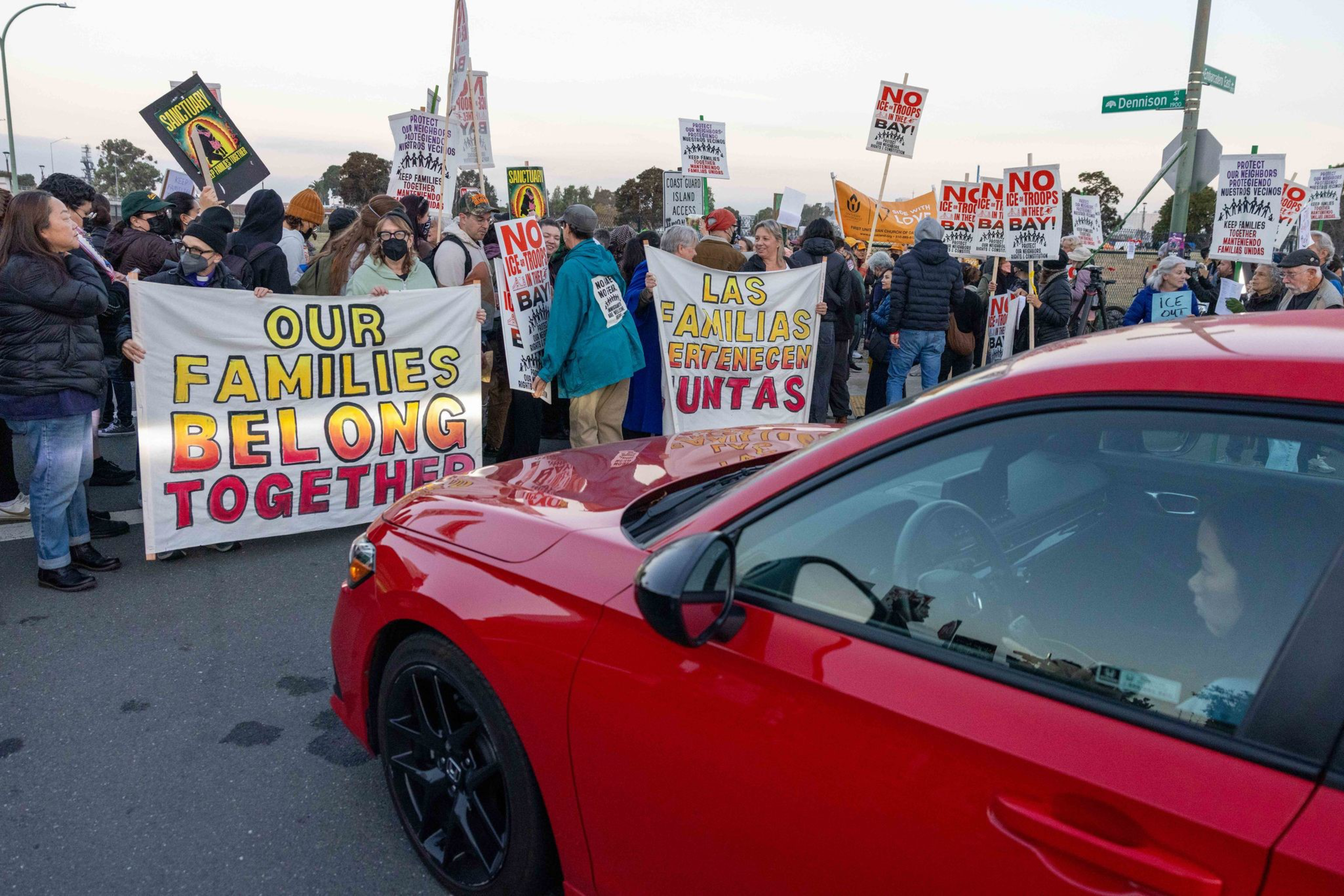 A crowd of people hold protest signs reading “Our Families Belong Together” and “Las Familias Pertenecen Juntas” blocking a street, with a red car nearby.