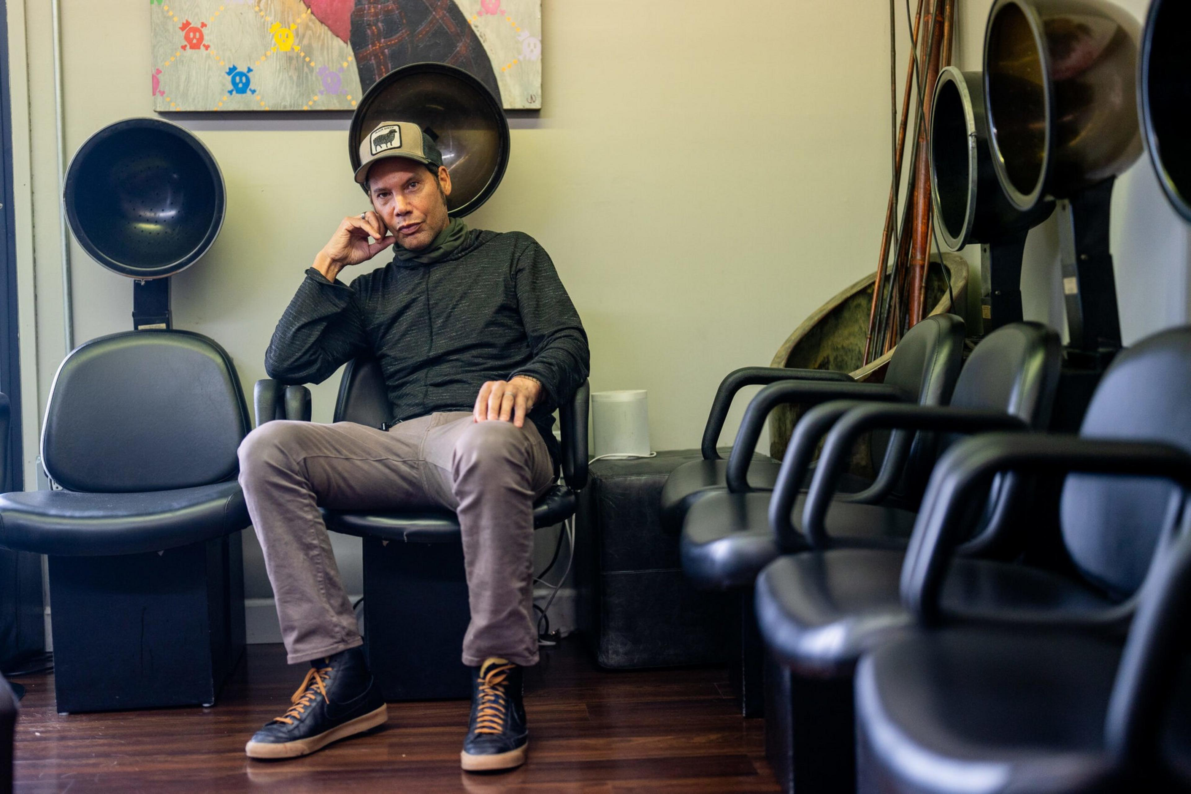 A man in a black jacket and cap sits relaxed in a salon chair between two hair drying stations in an empty room with wooden floors.