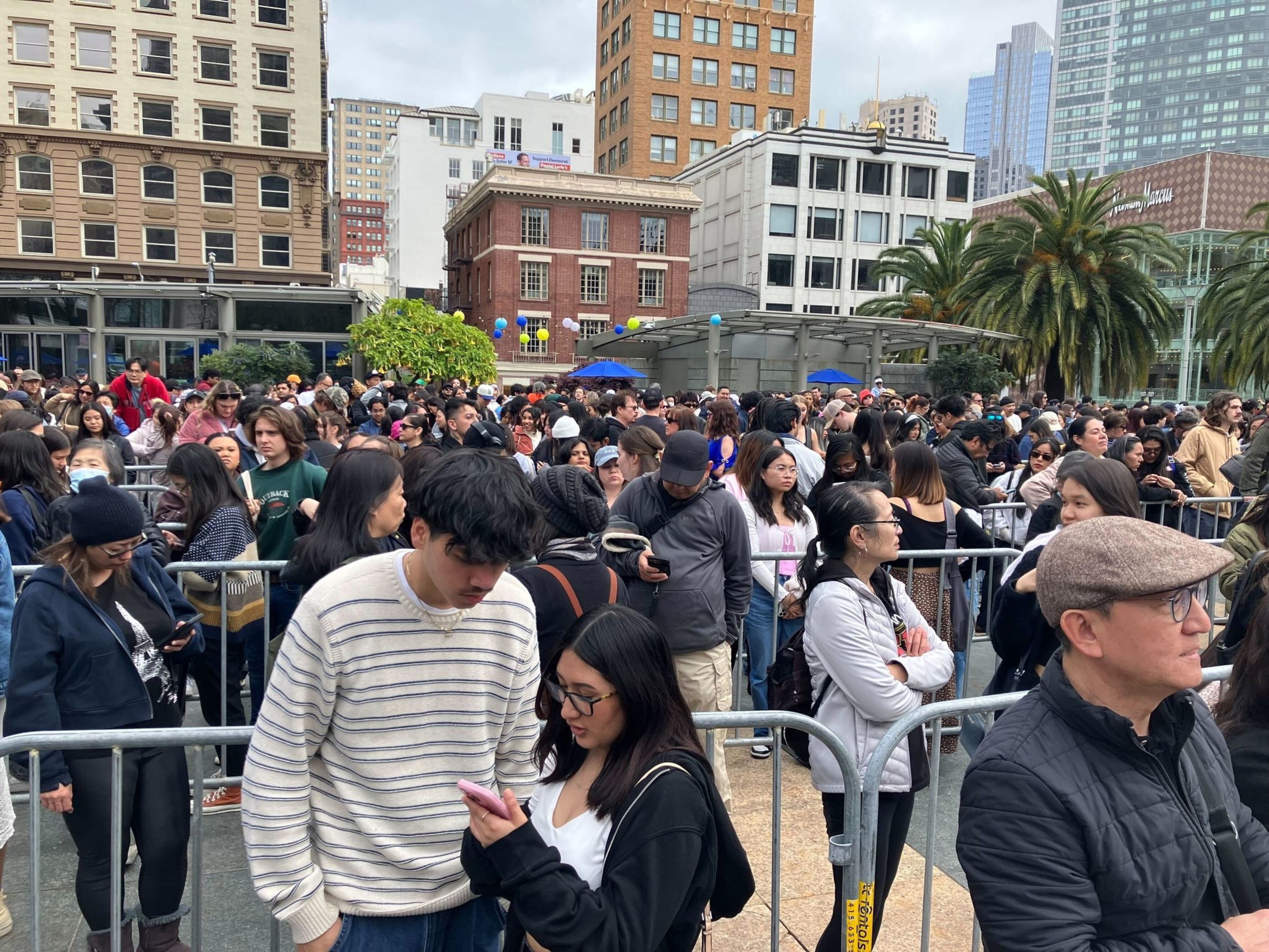 A crowded outdoor event with people queued up. There are buildings in the background and a few palm trees.
