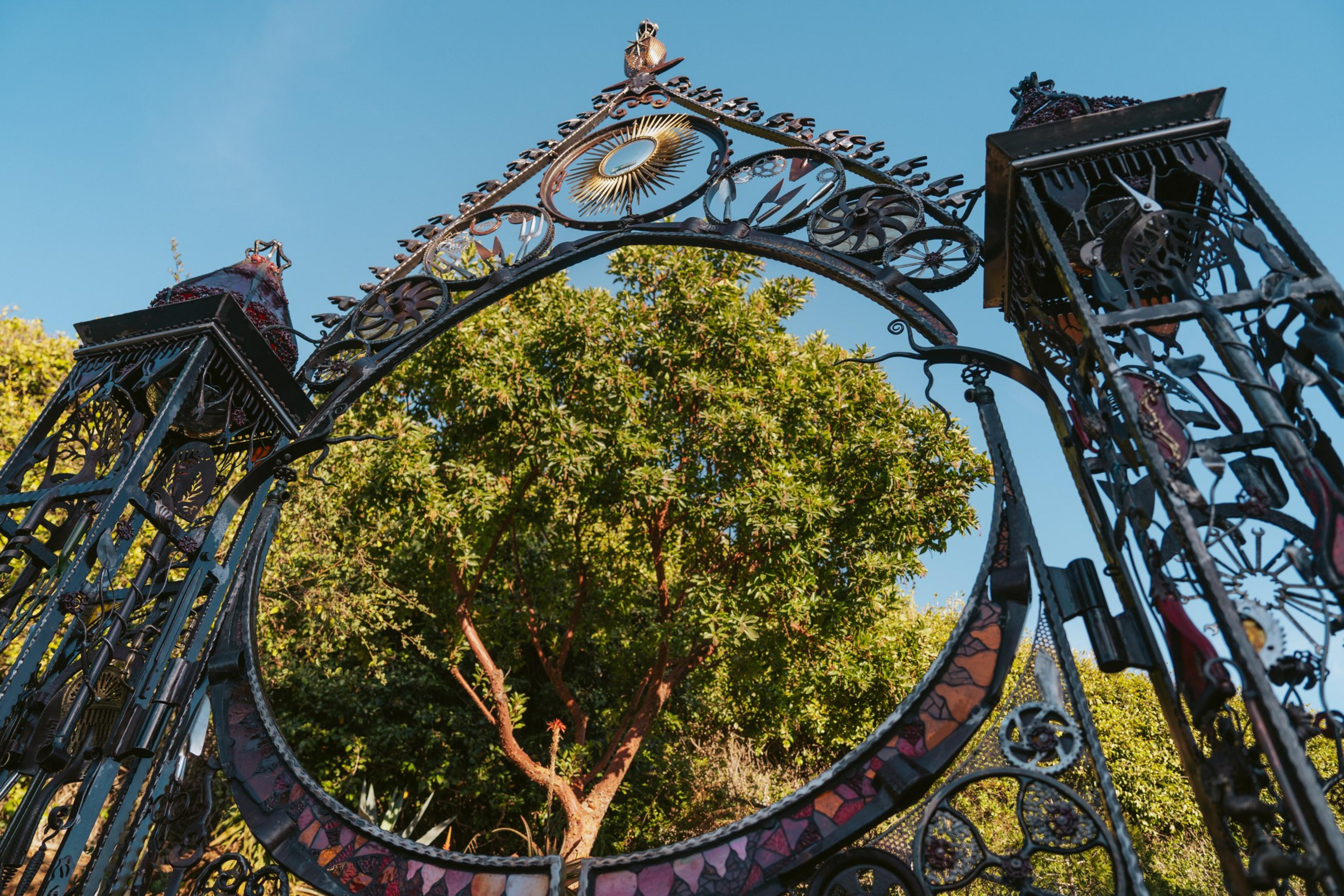 An ornate wrought iron gate with sun and gear designs frames a leafy tree, set against a clear blue sky on a sunny day.