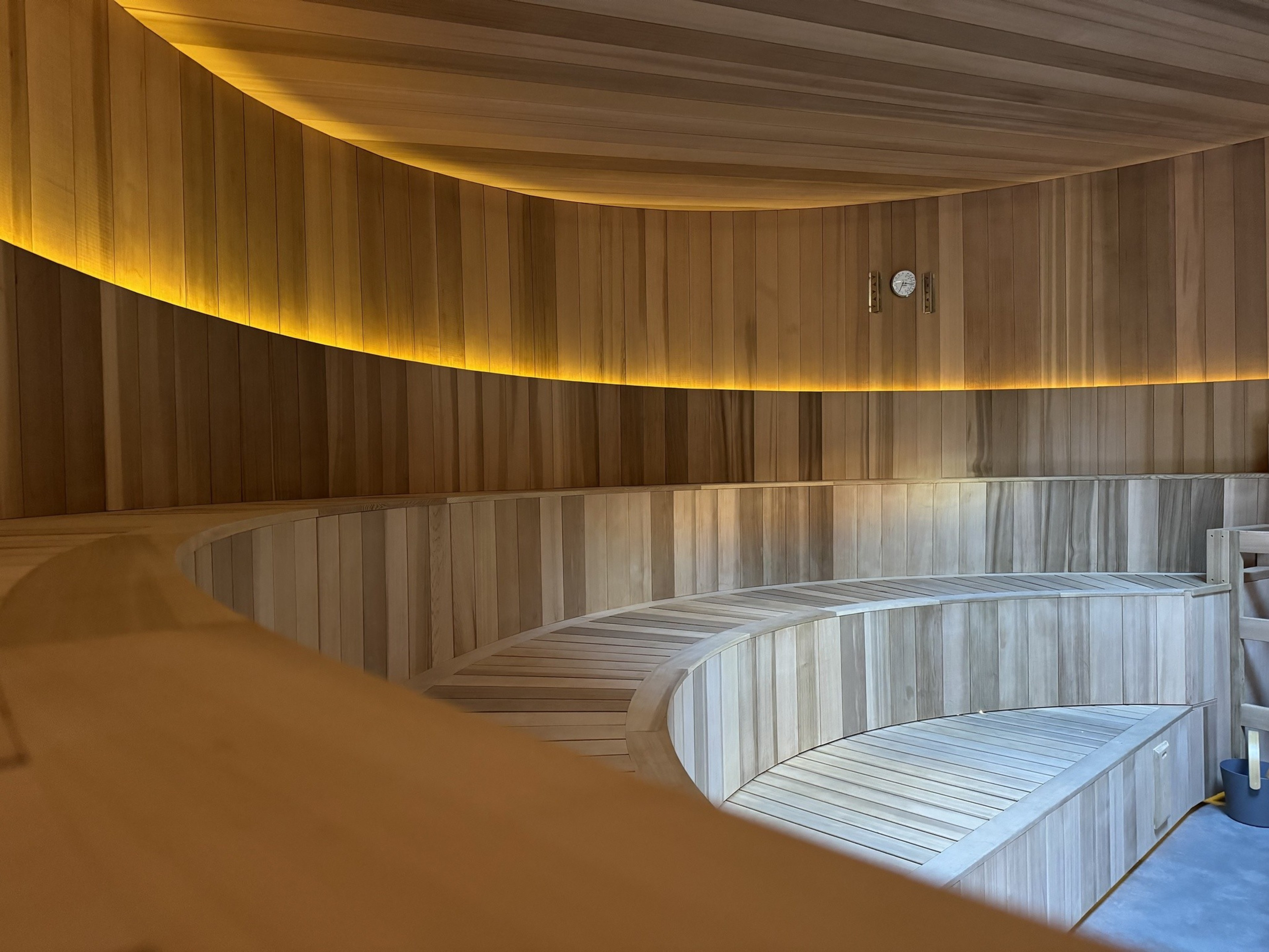 A wooden sauna with curved benches and warm, ambient lighting. A thermometer and hygrometer are mounted on the wall, adding to the cozy atmosphere.