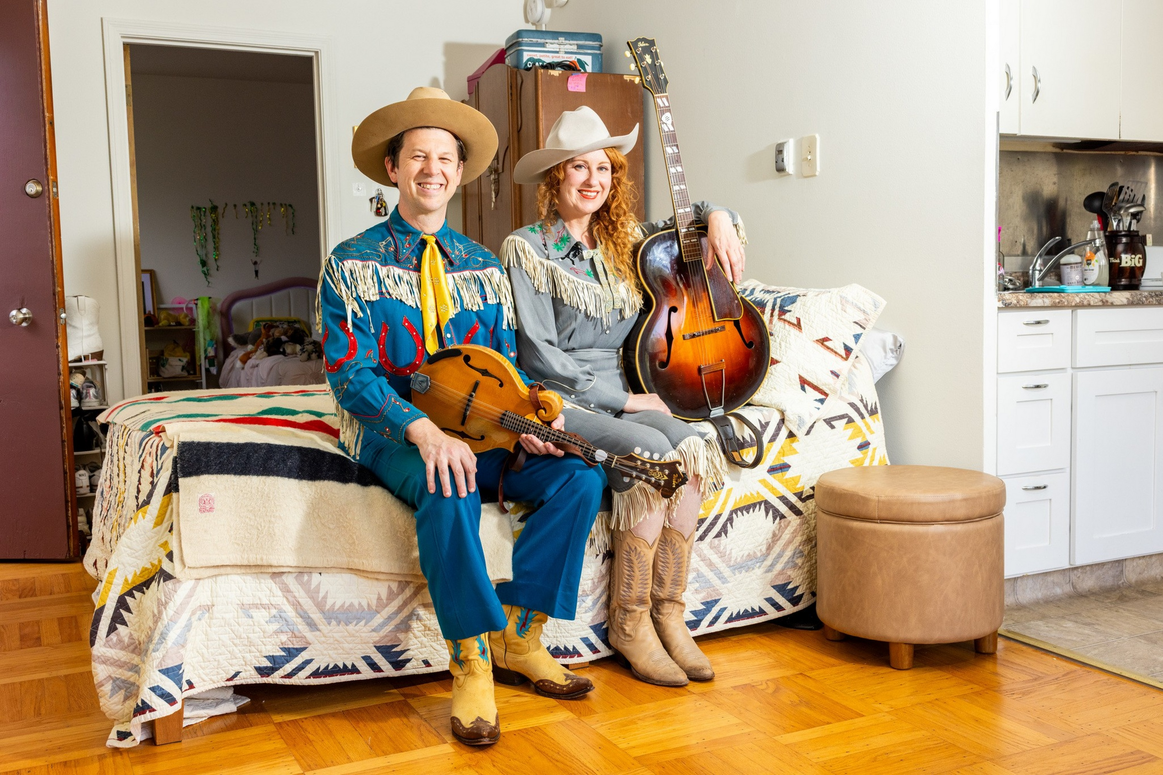 A man and woman in cowboy hats and fringed outfits sit on a patterned-covered couch, holding a mandolin and guitar in a cozy living room.
