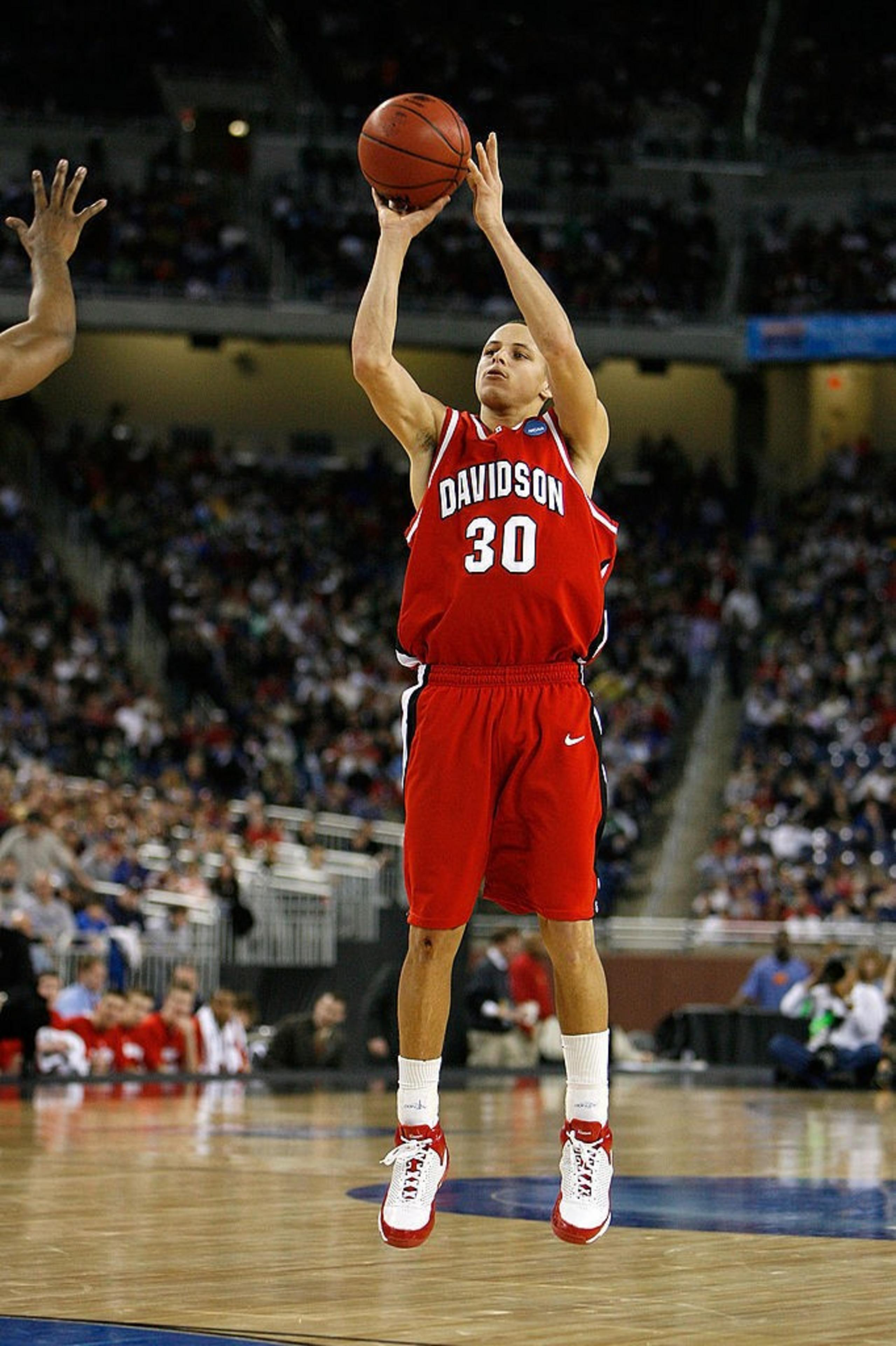 A basketball player in a red &quot;Davidson&quot; jersey, number 30, is mid-jump, shooting a basketball towards the hoop. The crowd watches in a stadium setting.