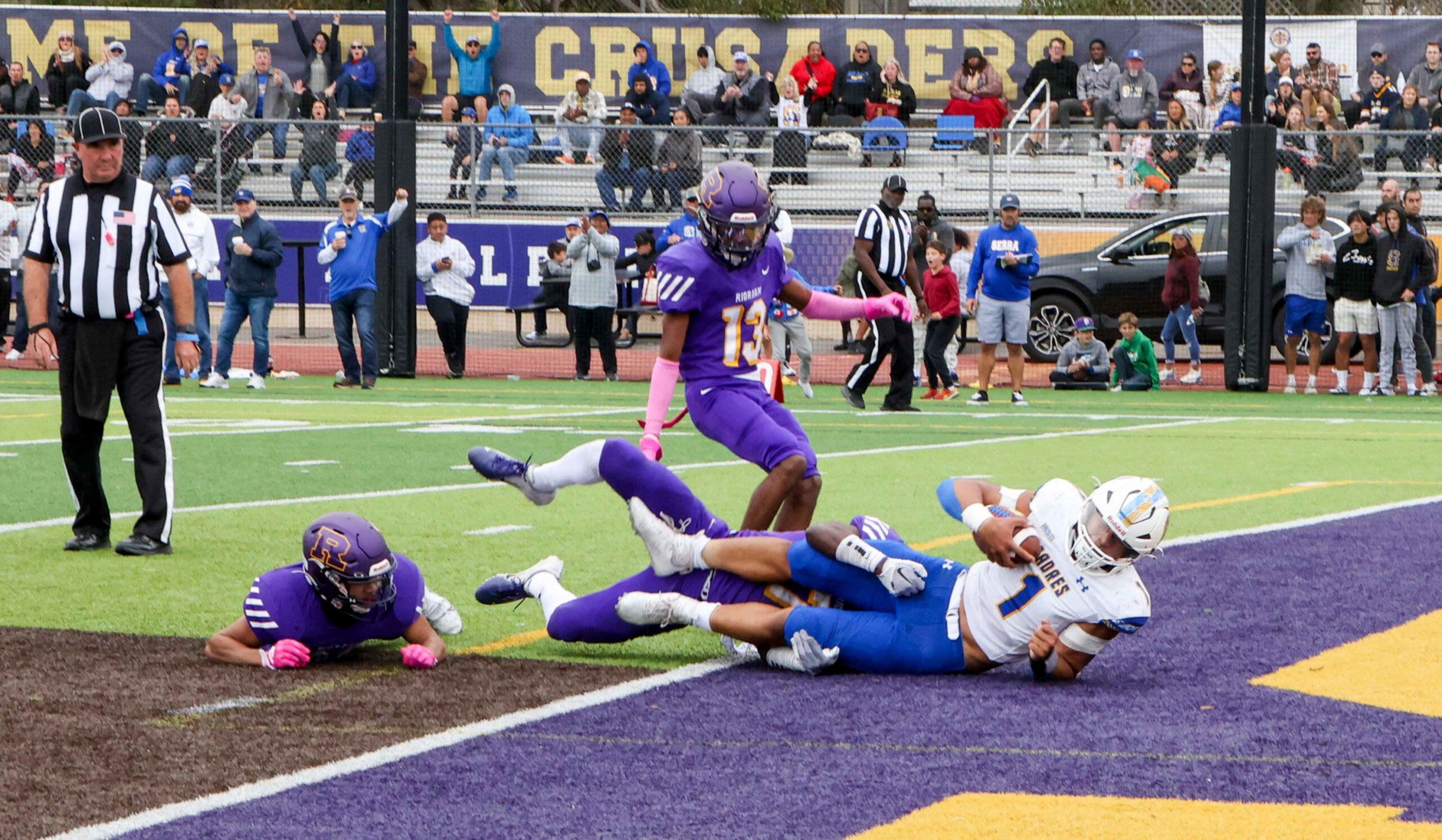 A football player in white and blue lies on the ground holding the ball, while two purple-clad defenders and a referee watch nearby on the field.