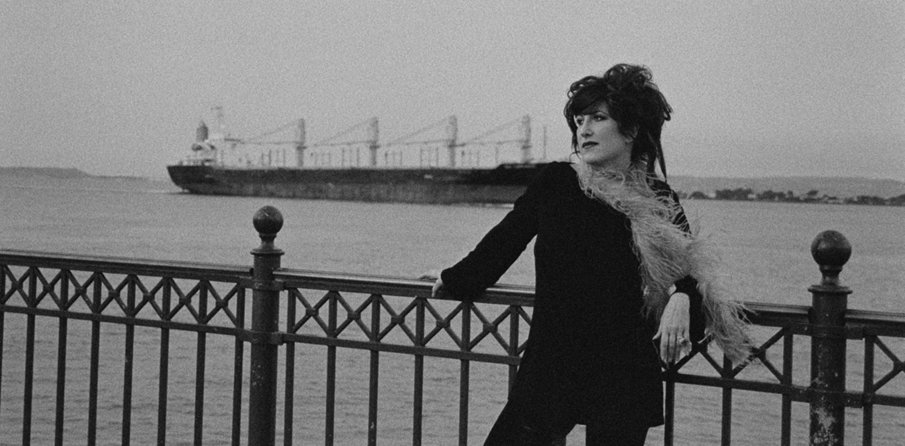 A woman in a dark outfit and feathered scarf leans on a railing by the water, with a large cargo ship in the distance behind her.