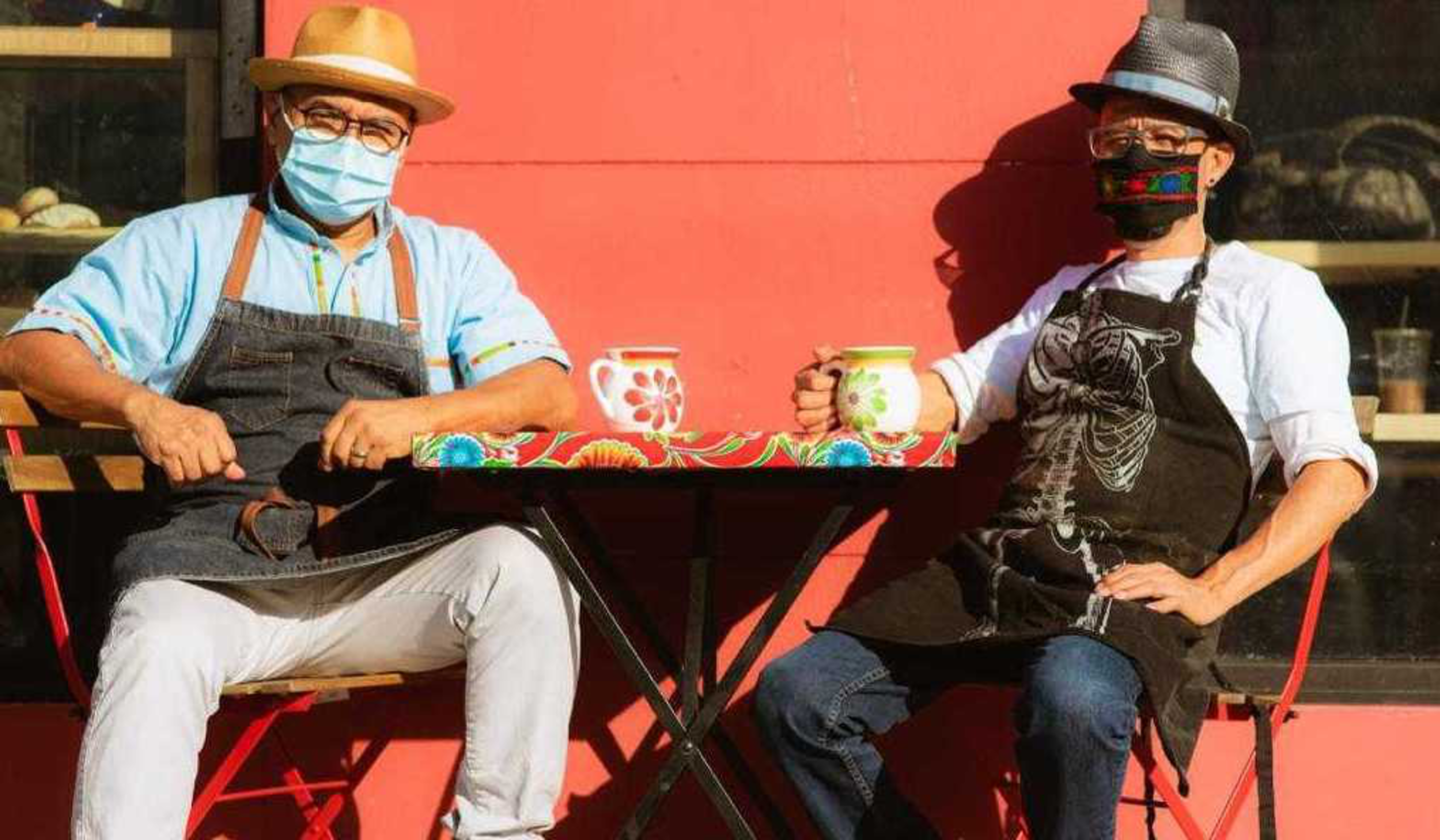Two people wearing hats, aprons, and face masks sit at a colorful table outdoors, each holding a decorated ceramic mug.