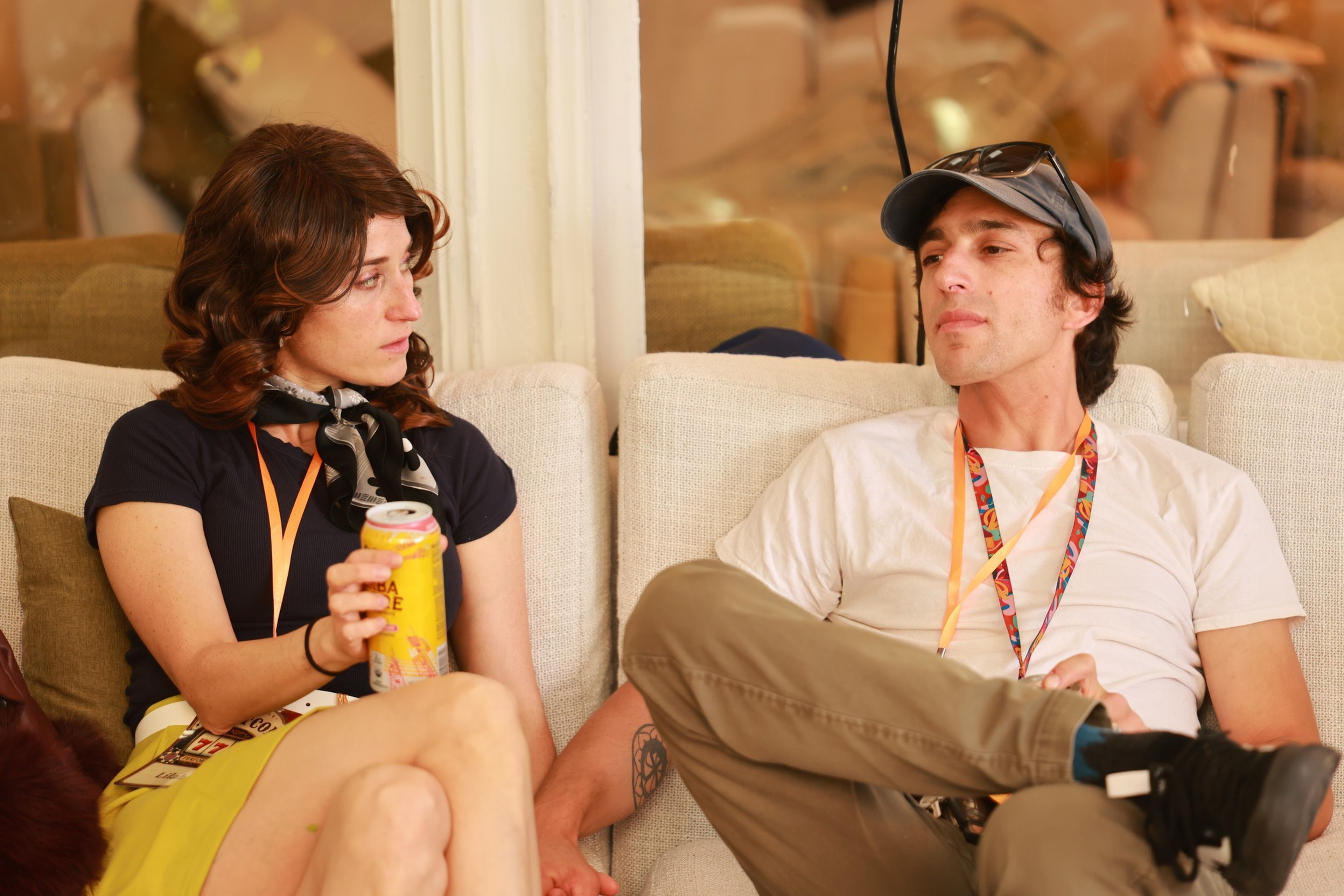 A woman with curly brown hair holds a yellow can while sitting next to a man in a white t-shirt and cap on a beige couch, both wearing lanyards.