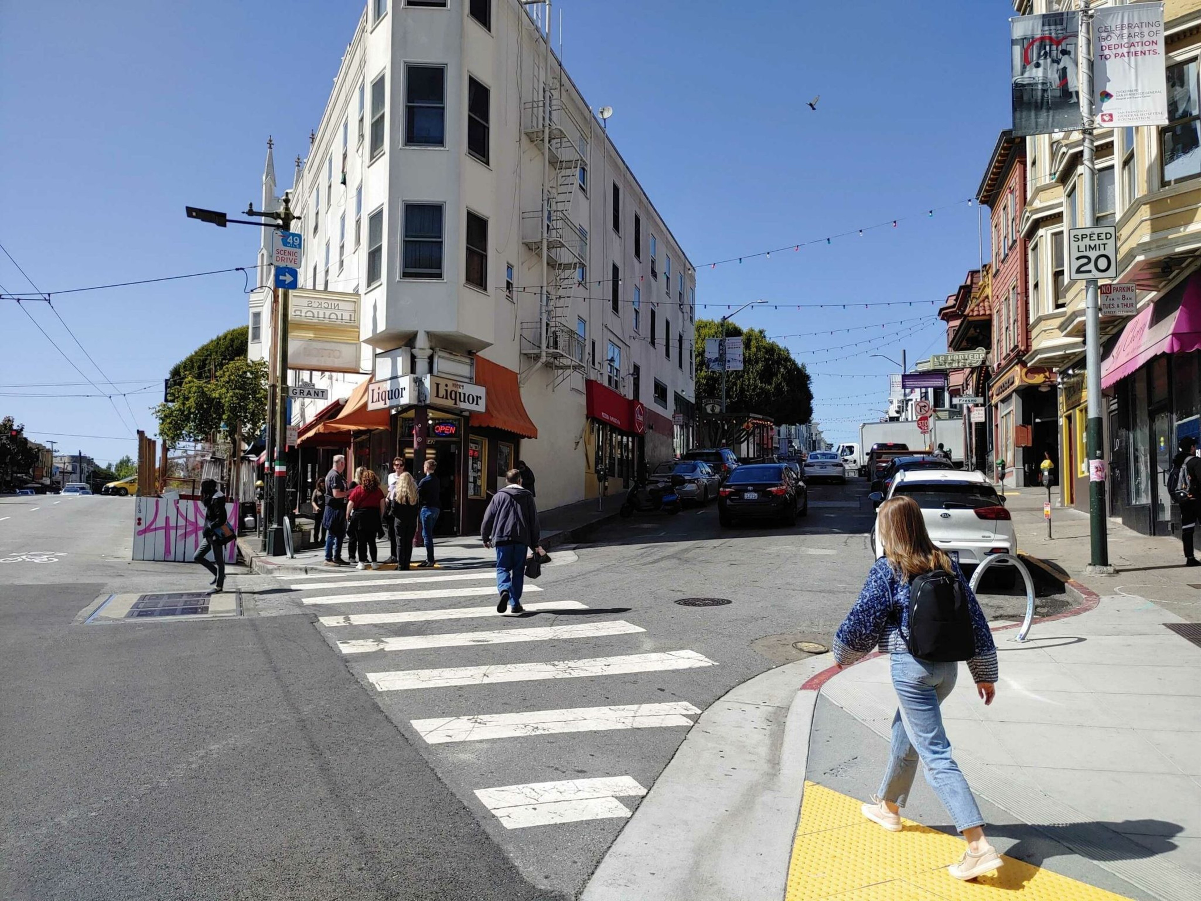 People cross streets at an intersection near a liquor store and other businesses on a sunny day with clear blue skies.