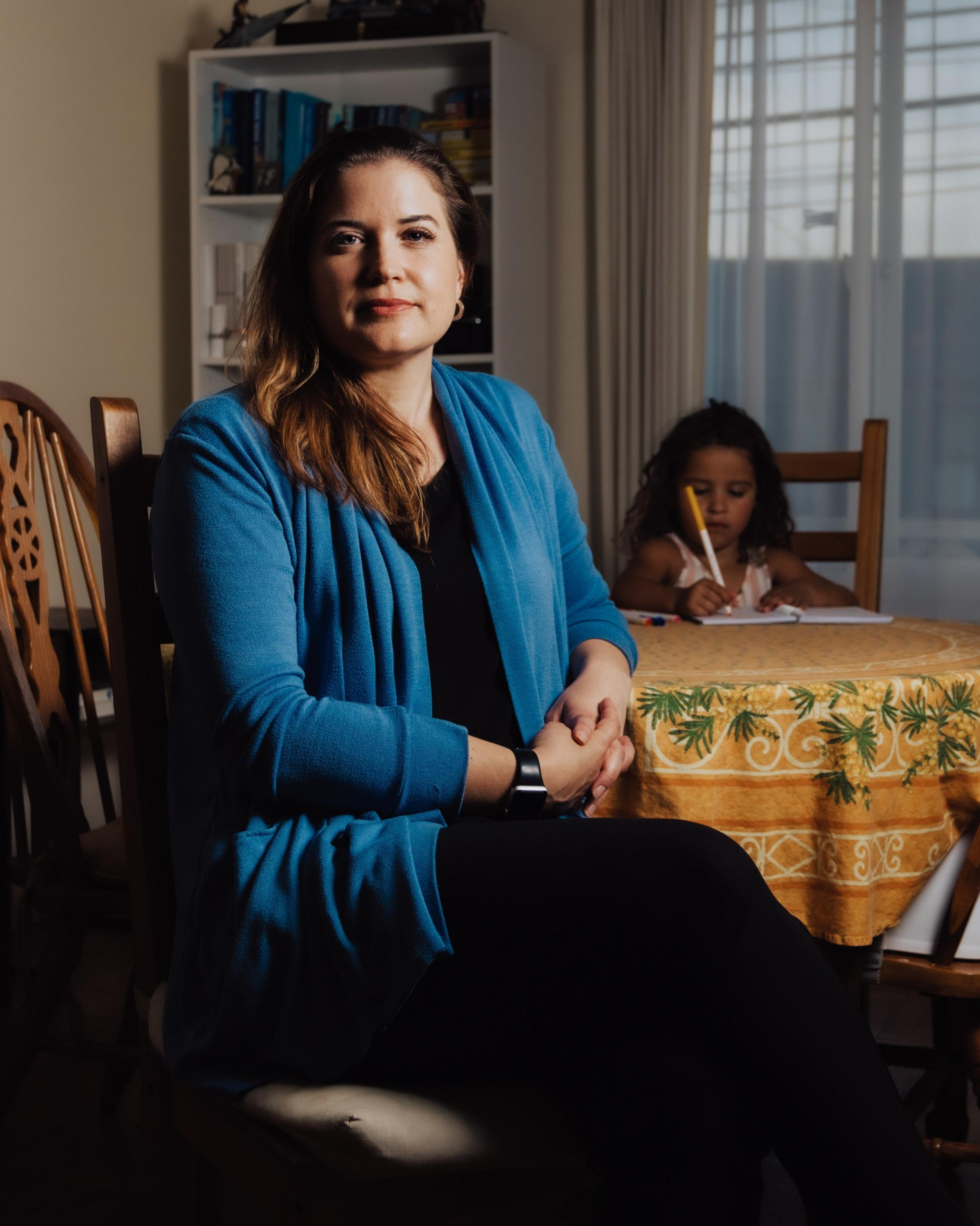 A woman in a blue sweater sits at a table with folded hands, while a child behind her colors on a paper. A bookshelf and window are in the background.