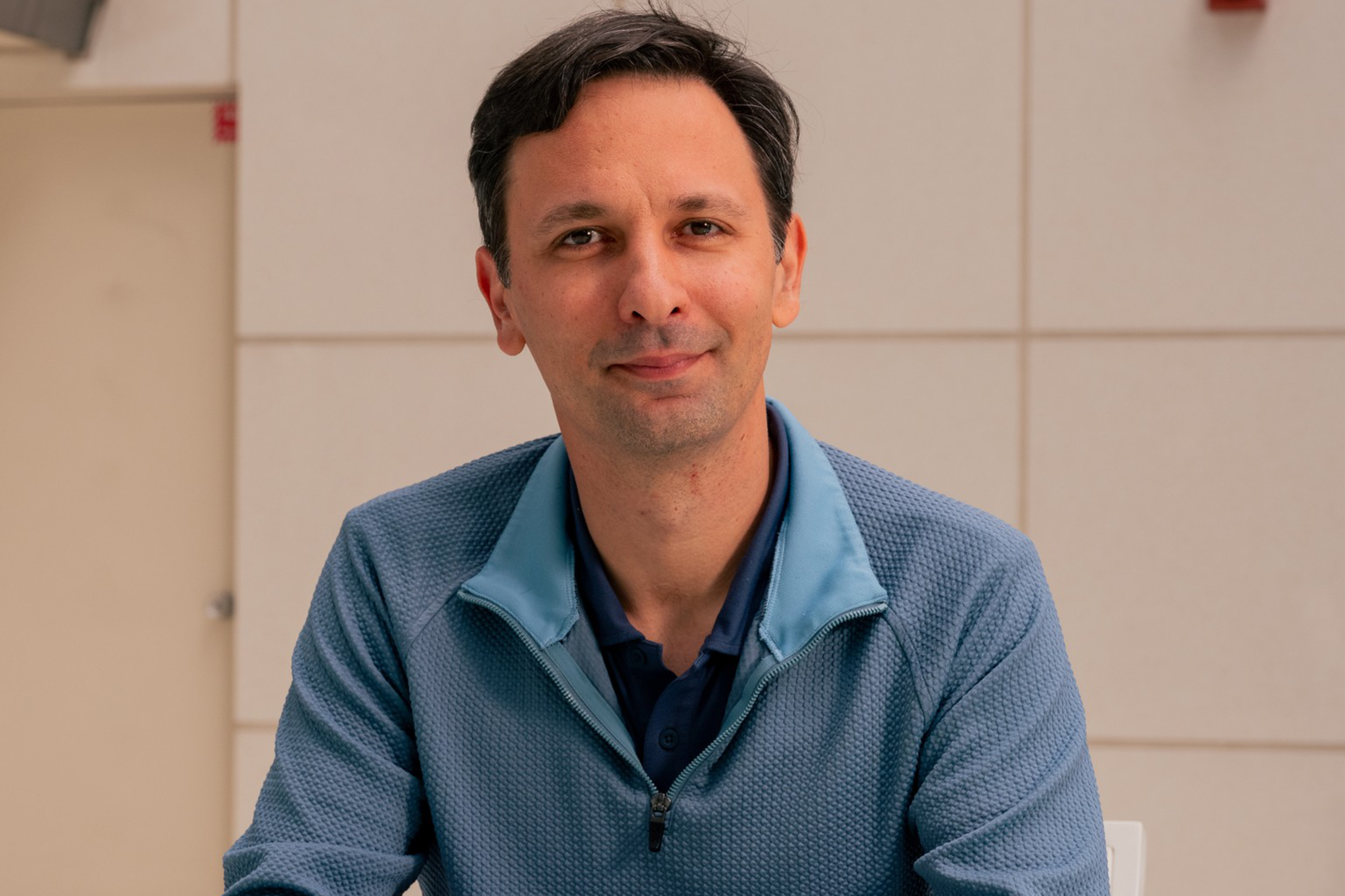A man with dark hair wears a blue zip-up sweater over a dark shirt, sitting at a glass table with hands clasped, against a tiled beige wall.