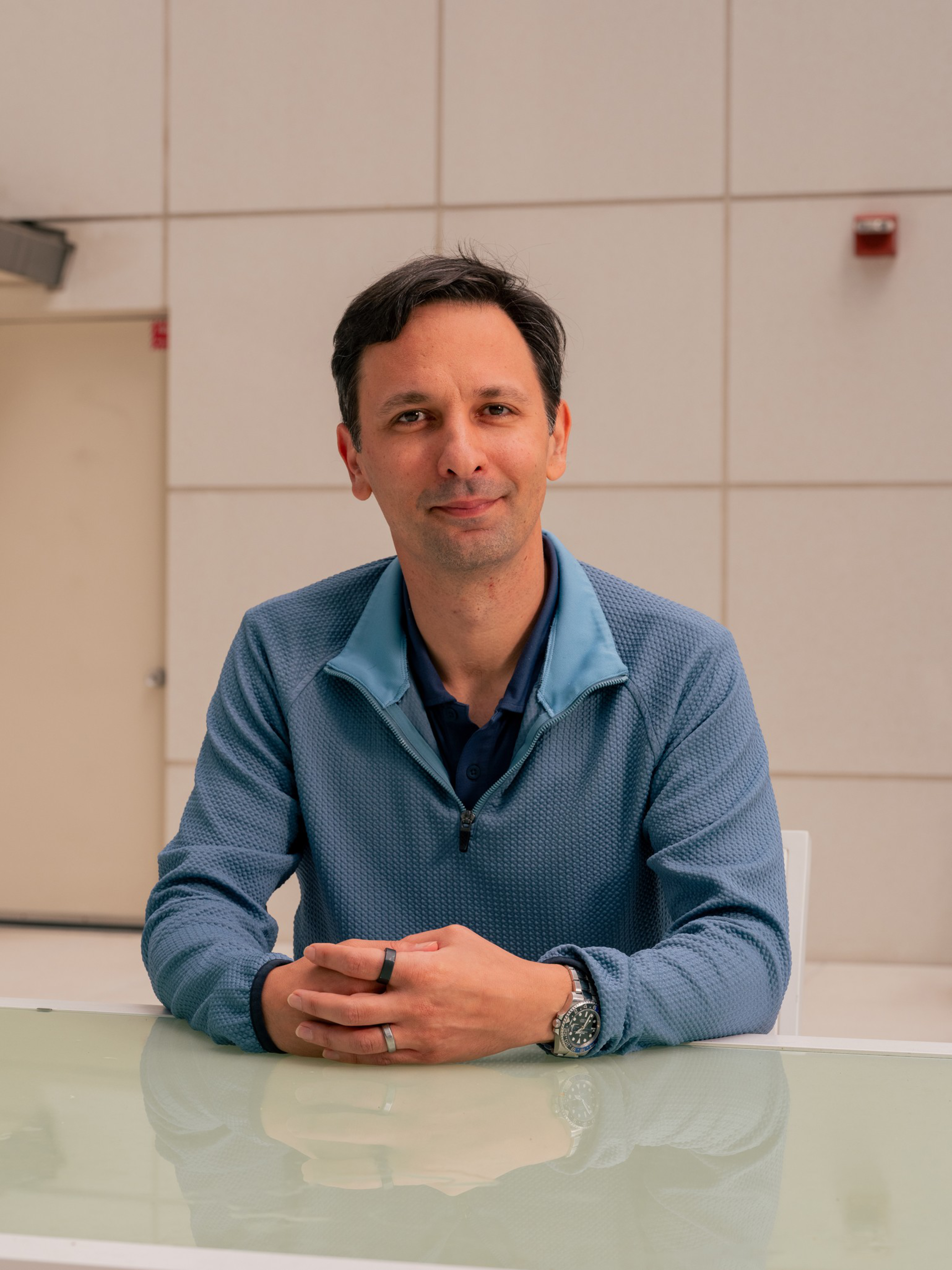 A man with dark hair wears a blue zip-up sweater over a dark shirt, sitting at a glass table with hands clasped, against a tiled beige wall.