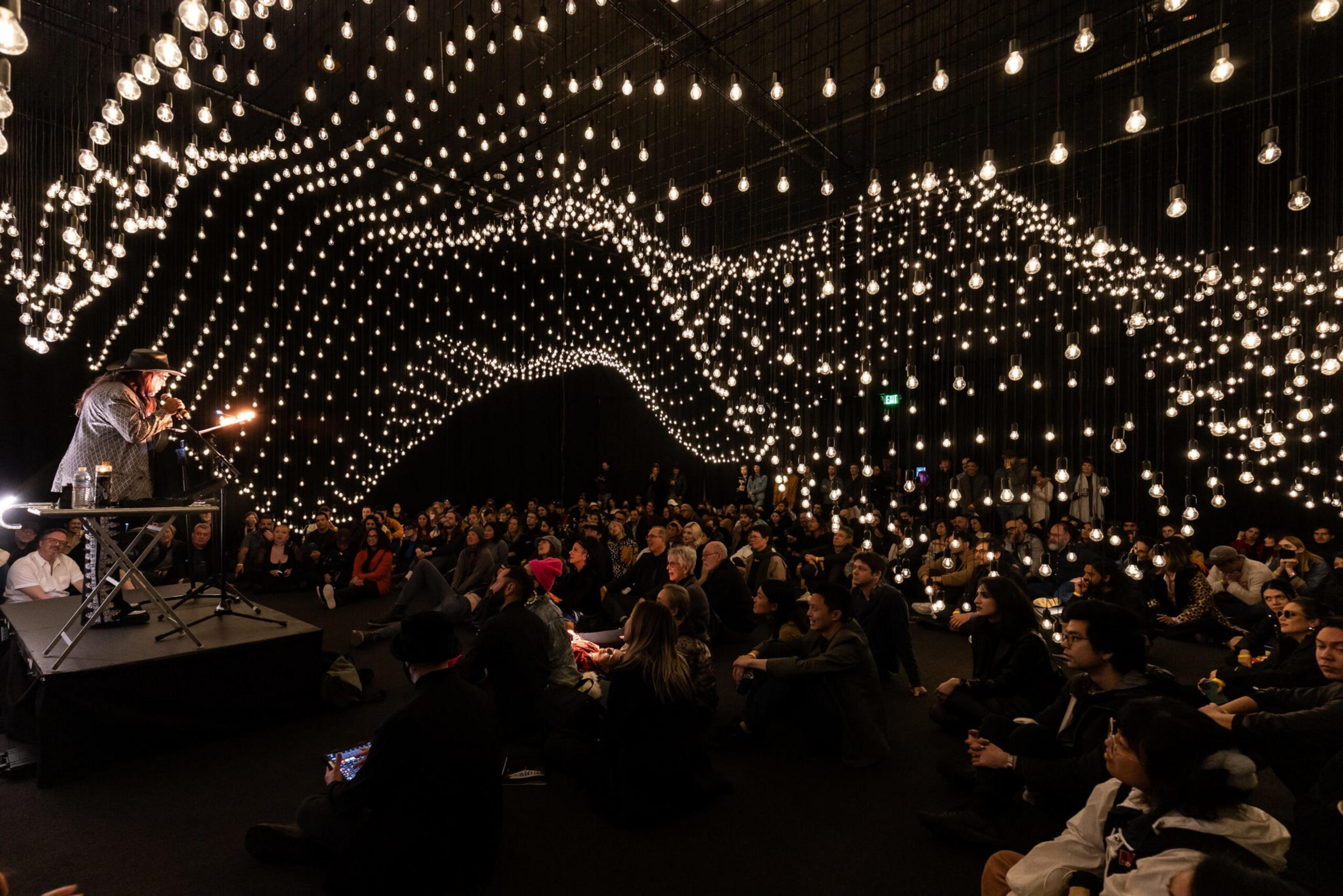 A person speaks into a microphone on a small stage while hundreds of hanging light bulbs create waves above a seated audience in a dark room.