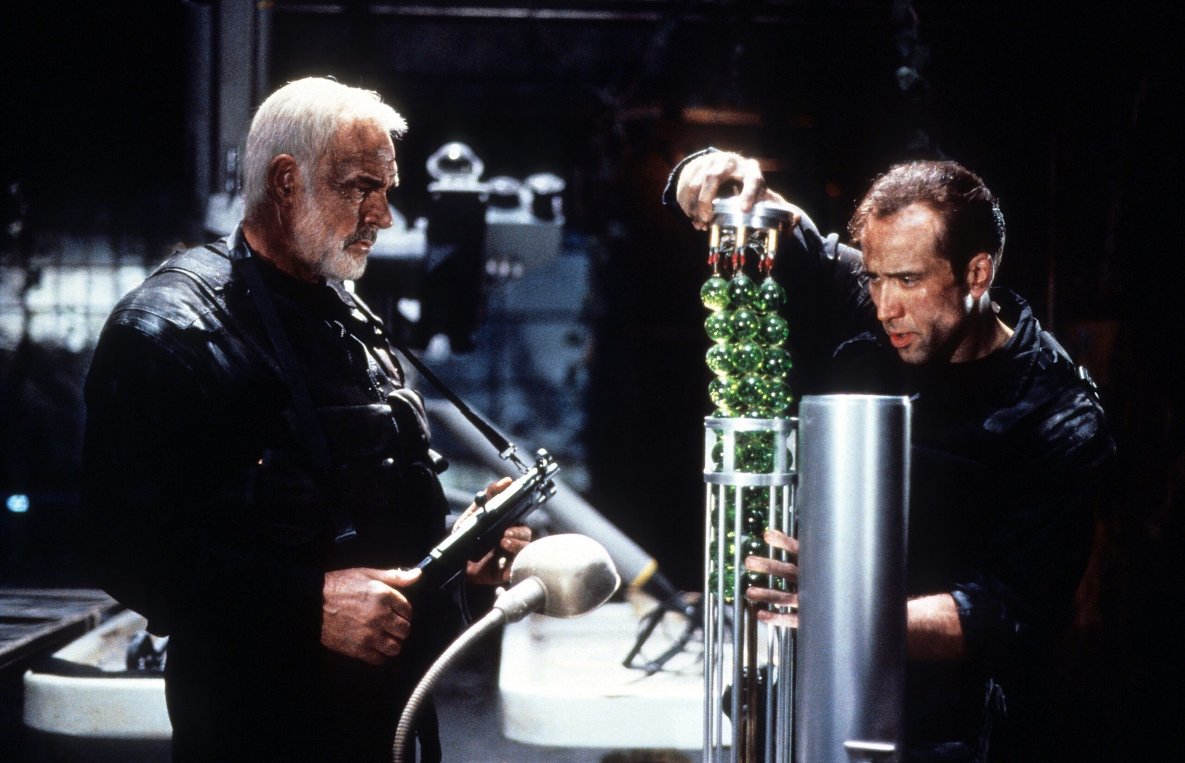 An older man with gray hair and a beard holds a gun while a younger man inspects a tall device filled with green spheres. Both wear dark clothing.