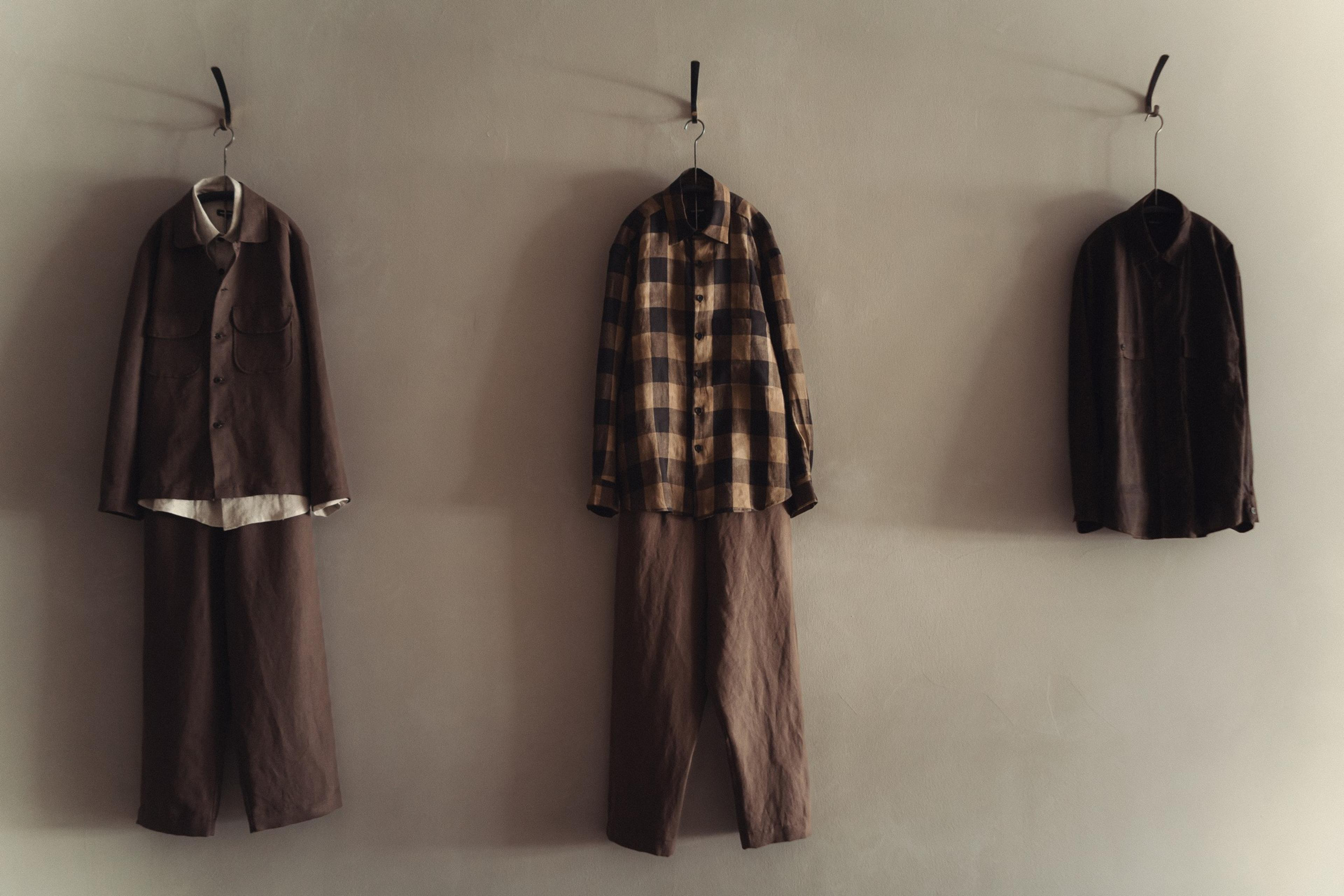 Three outfits hang on a wall: a brown jacket and pants, a checkered shirt with brown pants, and a dark button-up shirt.