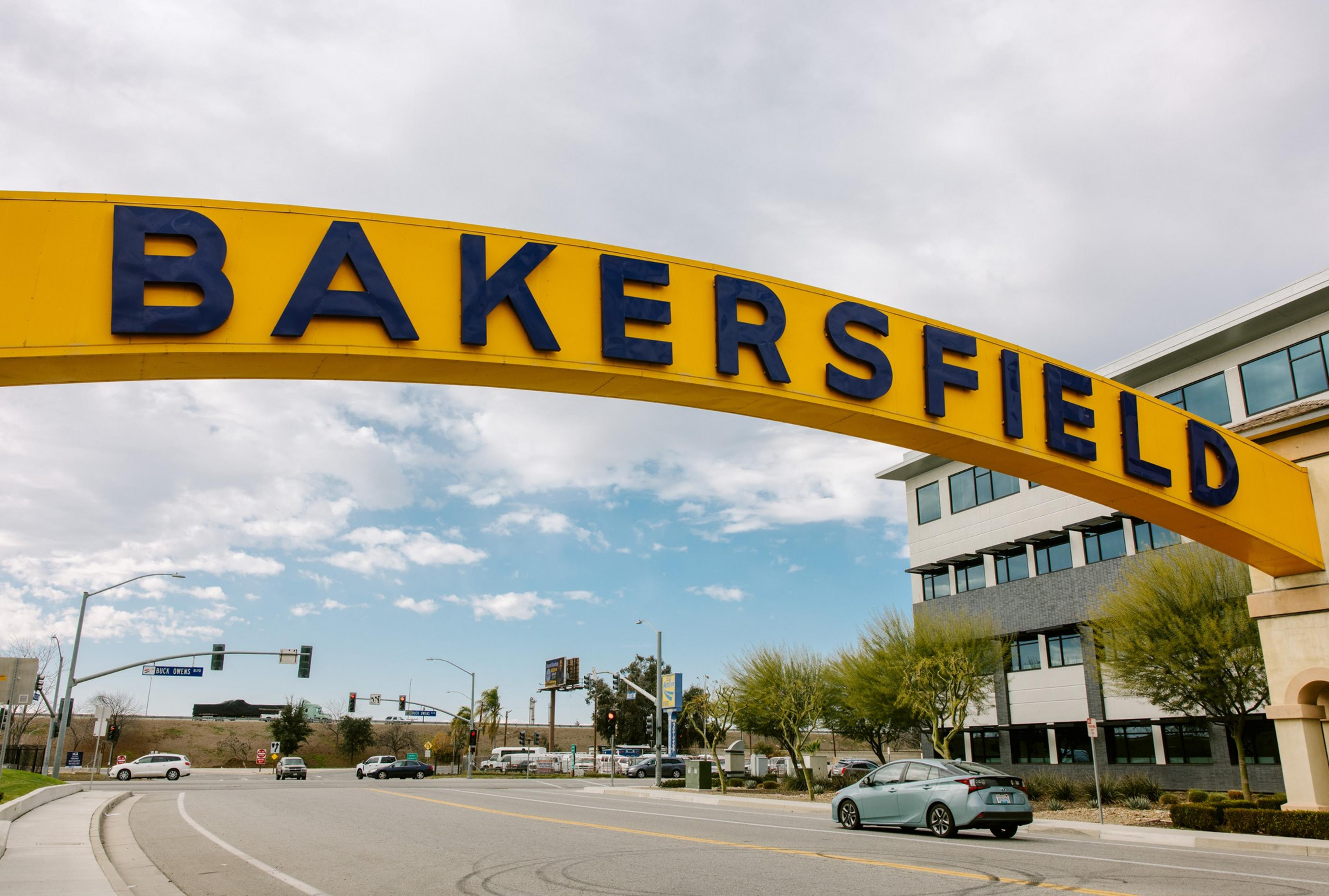 A large yellow arch with &quot;Bakersfield&quot; in bold blue letters spans over a street, with buildings and trees lining the road under a partly cloudy sky.