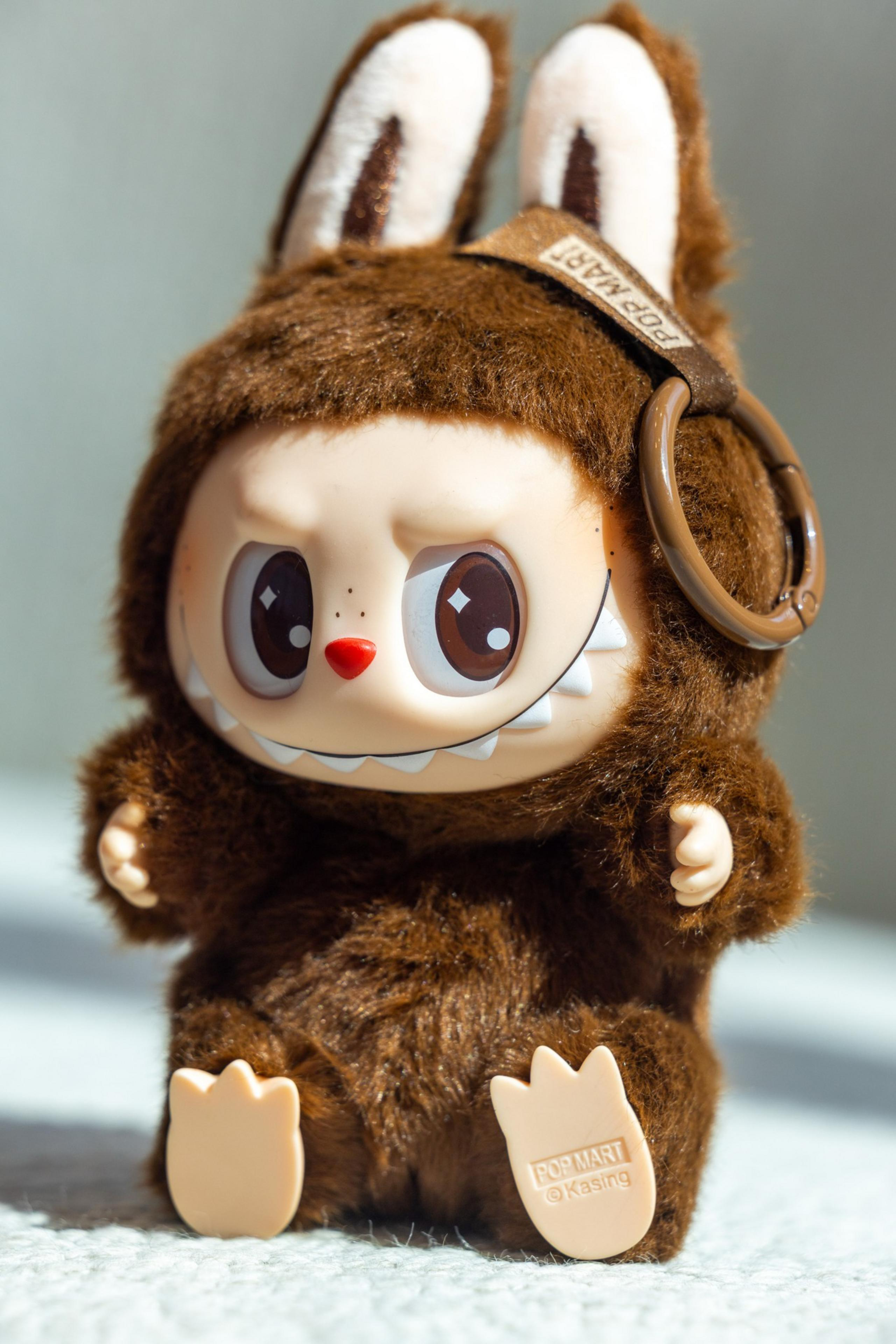 The image shows a cute toy figure with a big head, wearing a fuzzy brown costume with tall ears. It has large eyes, a small red nose, and a mischievous grin.