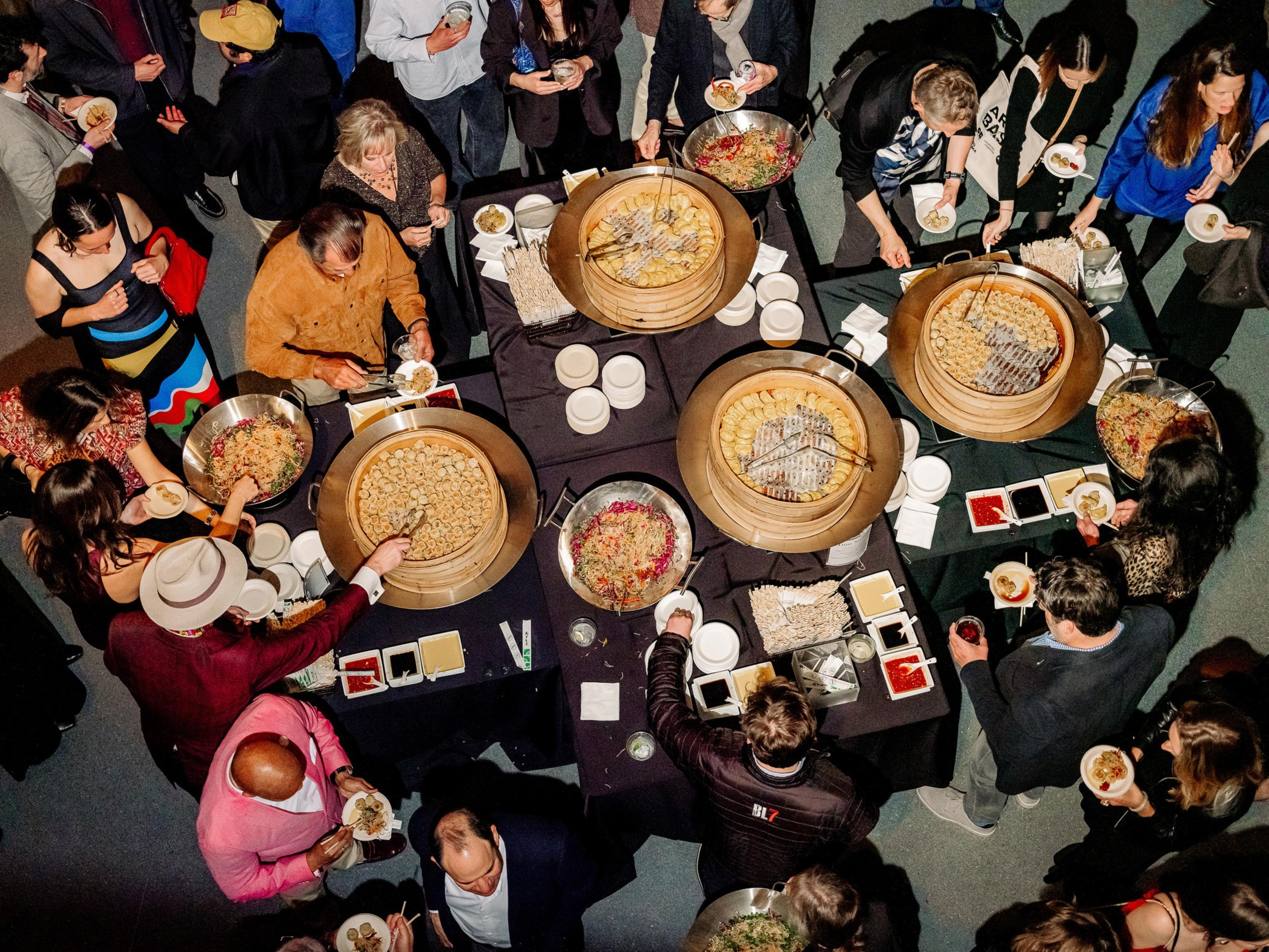 People gather around a buffet table with large trays of food, picking items onto small plates. The scene is lively, with various dishes and condiments.