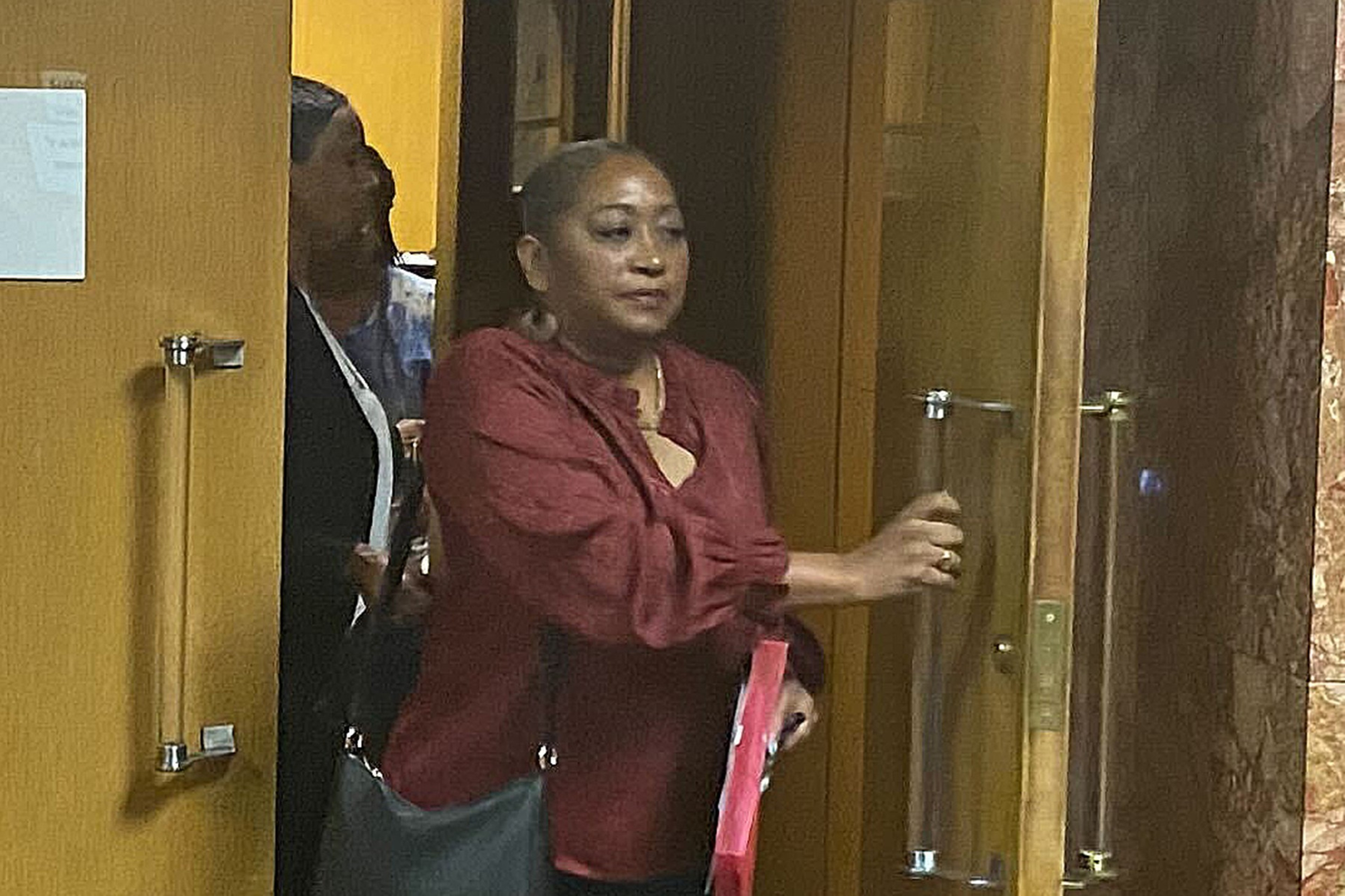 A person in a red blouse is opening a glass door, with another person peeking from behind a yellow door.