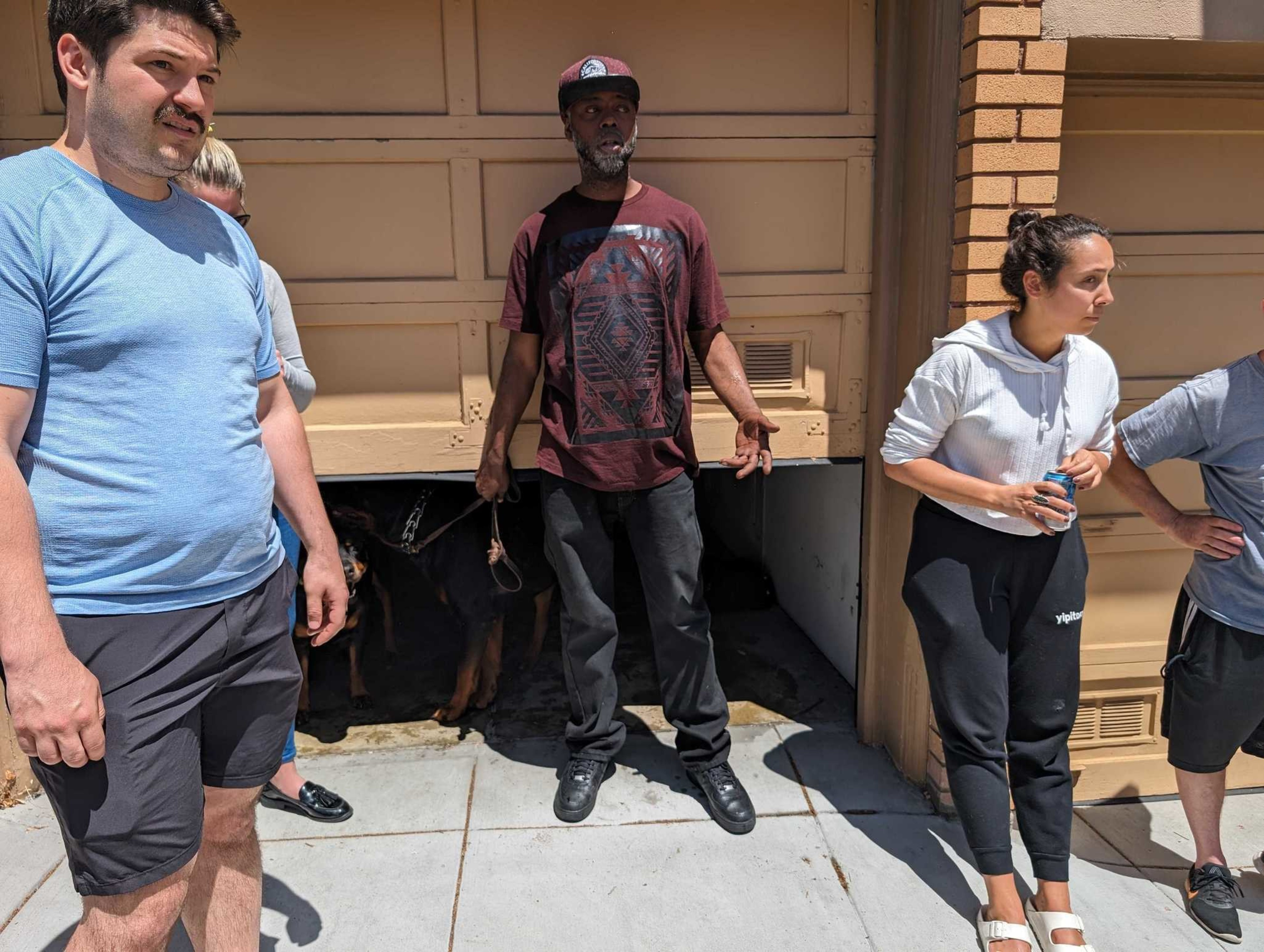Four adults stand in the sun by a building’s side, showing various casual poses and expressions, accompanied by a hidden dog on a leash.