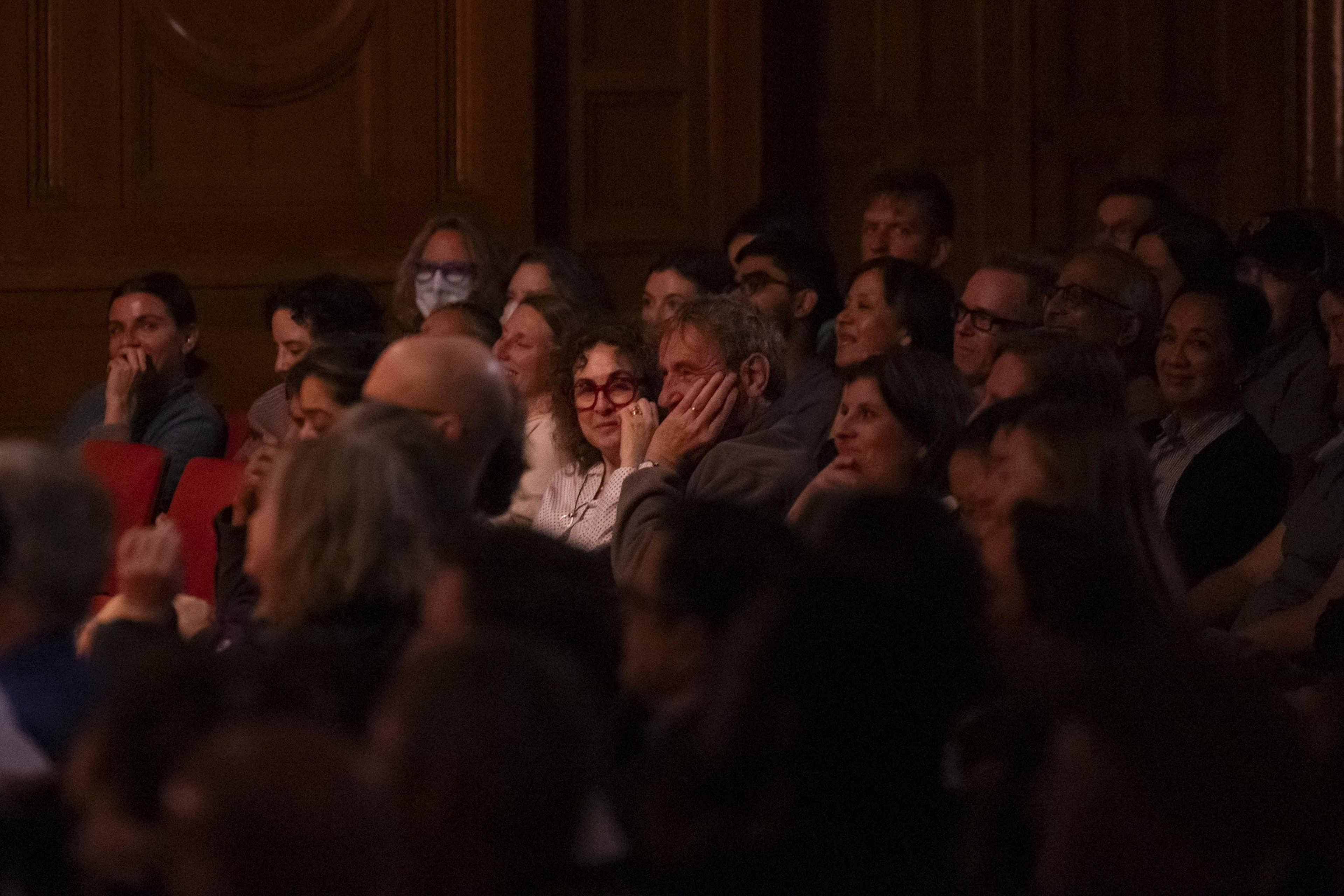 A dimly lit audience is seen, with people attentively watching something off-camera. One person in the center wears red glasses, smiling gently.