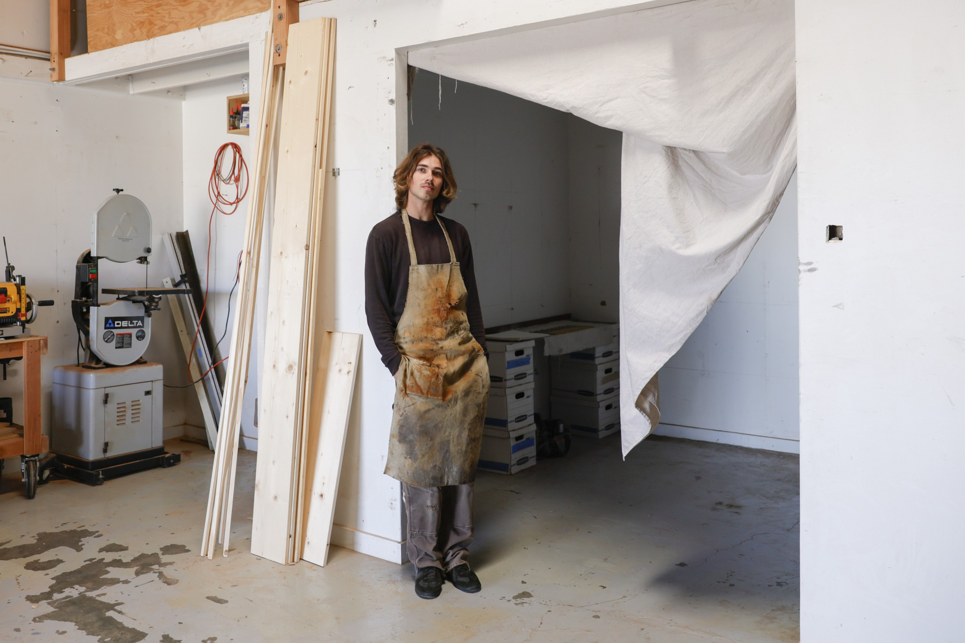 A person wearing a stained apron stands with hands in pockets in a workshop with wood planks, machines, and a partially covered open doorway.