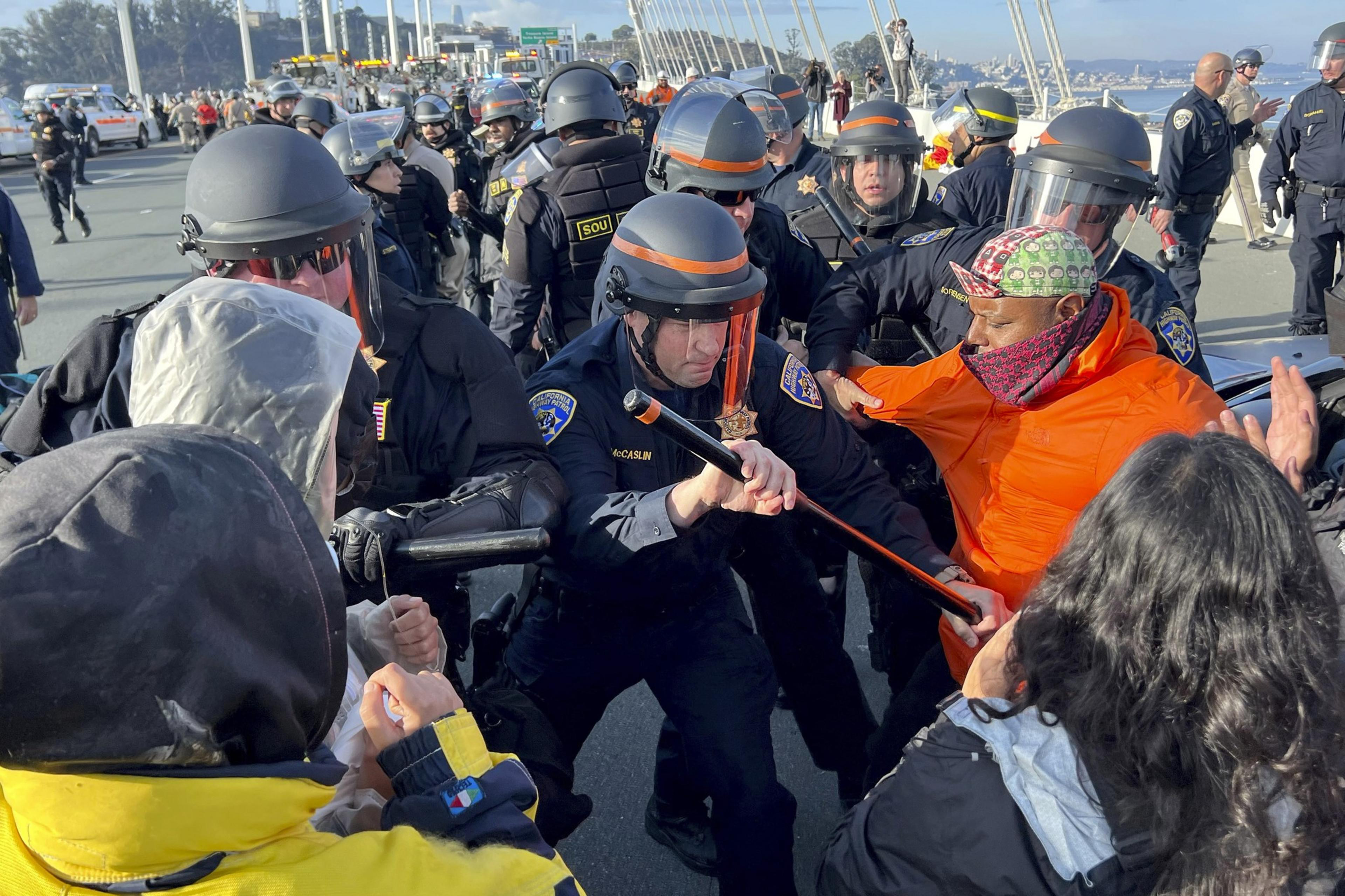 A large group of SFPD officers wearing riot gear use batons on protesters during a protest on the Bay Bridge.
