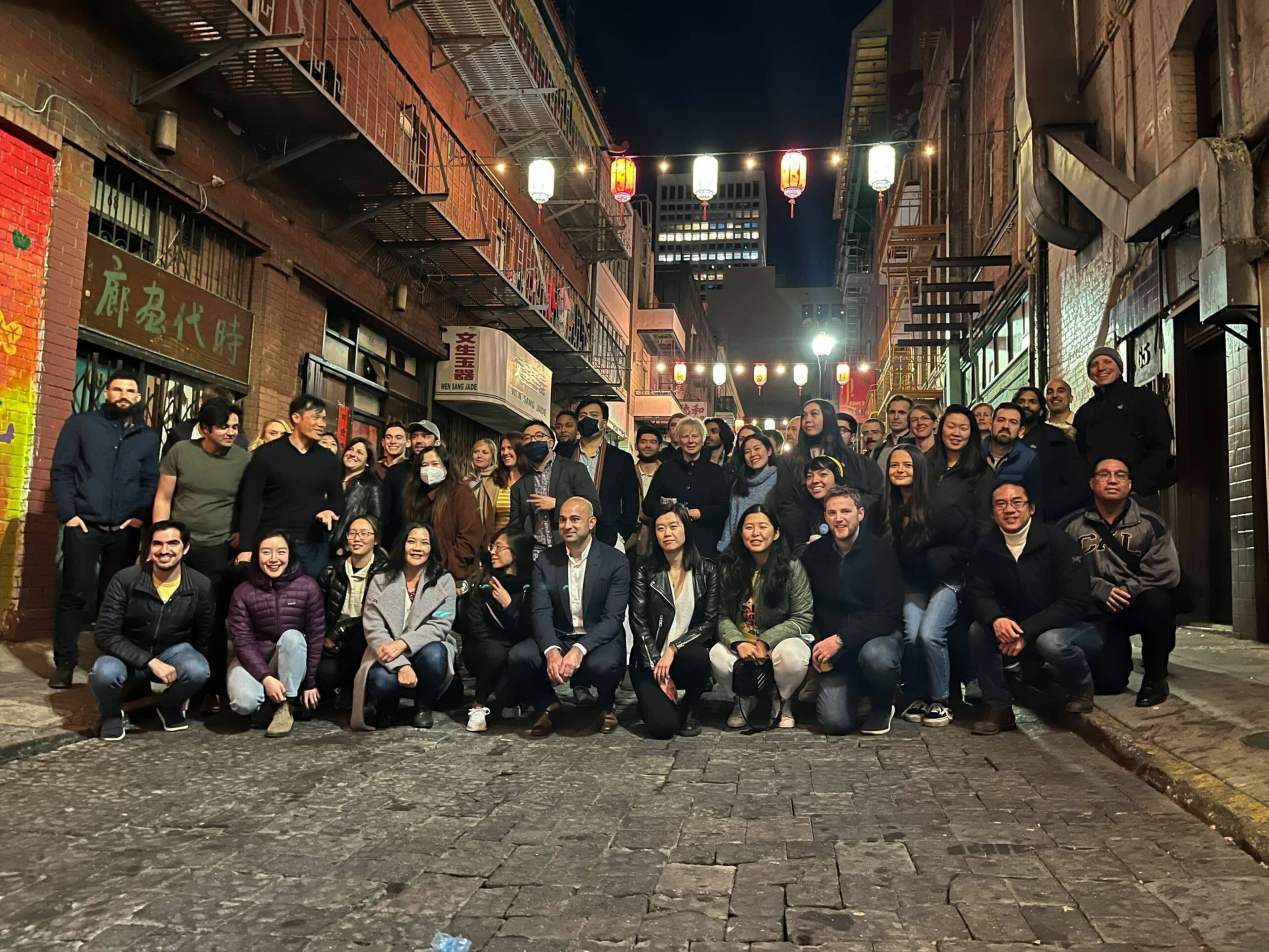 A large group of people poses together on a cobblestone street at night, with illuminated lanterns hanging above and brick buildings on both sides.