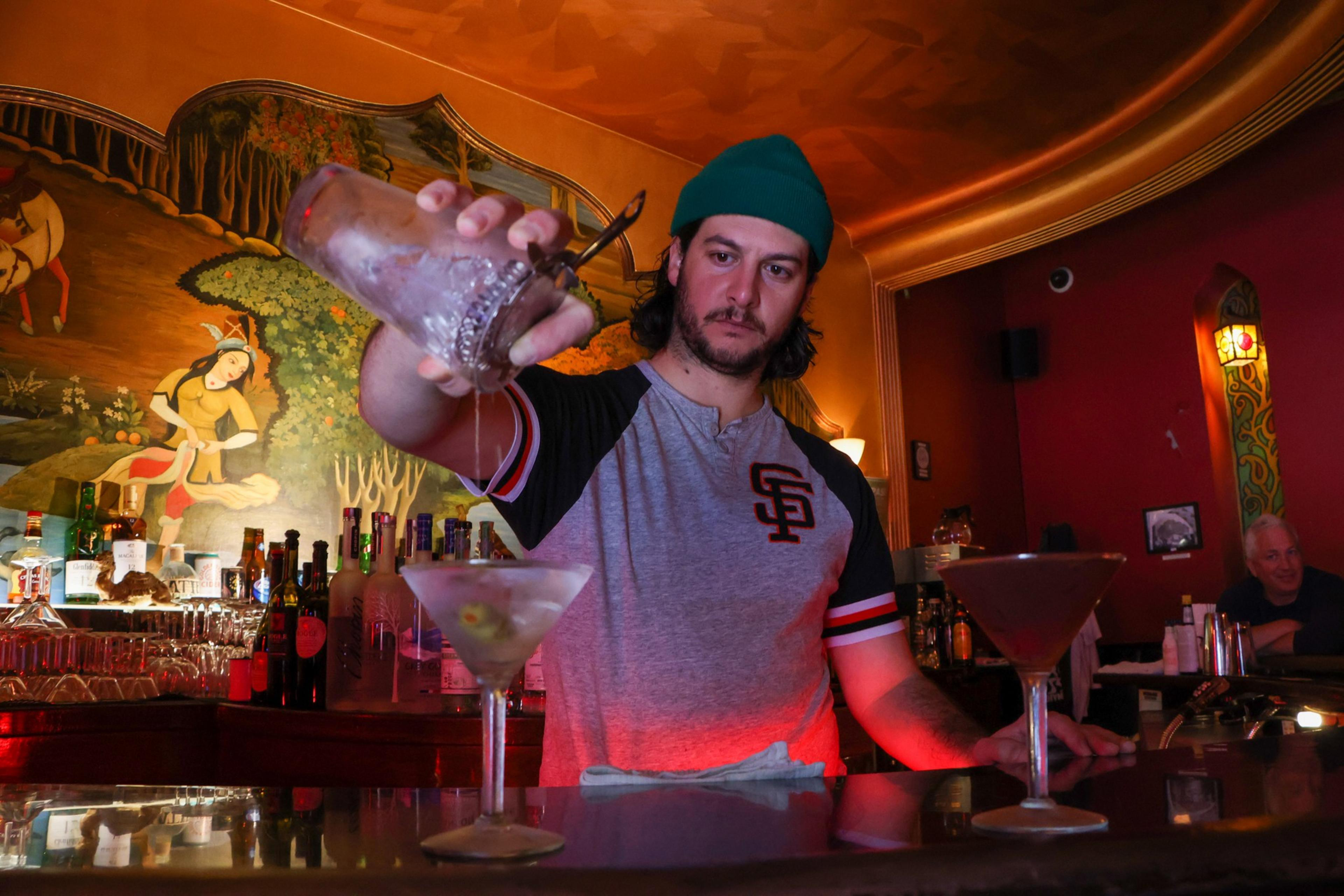 A bartender wearing a teal beanie and gray SF shirt pours a drink from a shaker into one of two martini glasses on the bar counter.